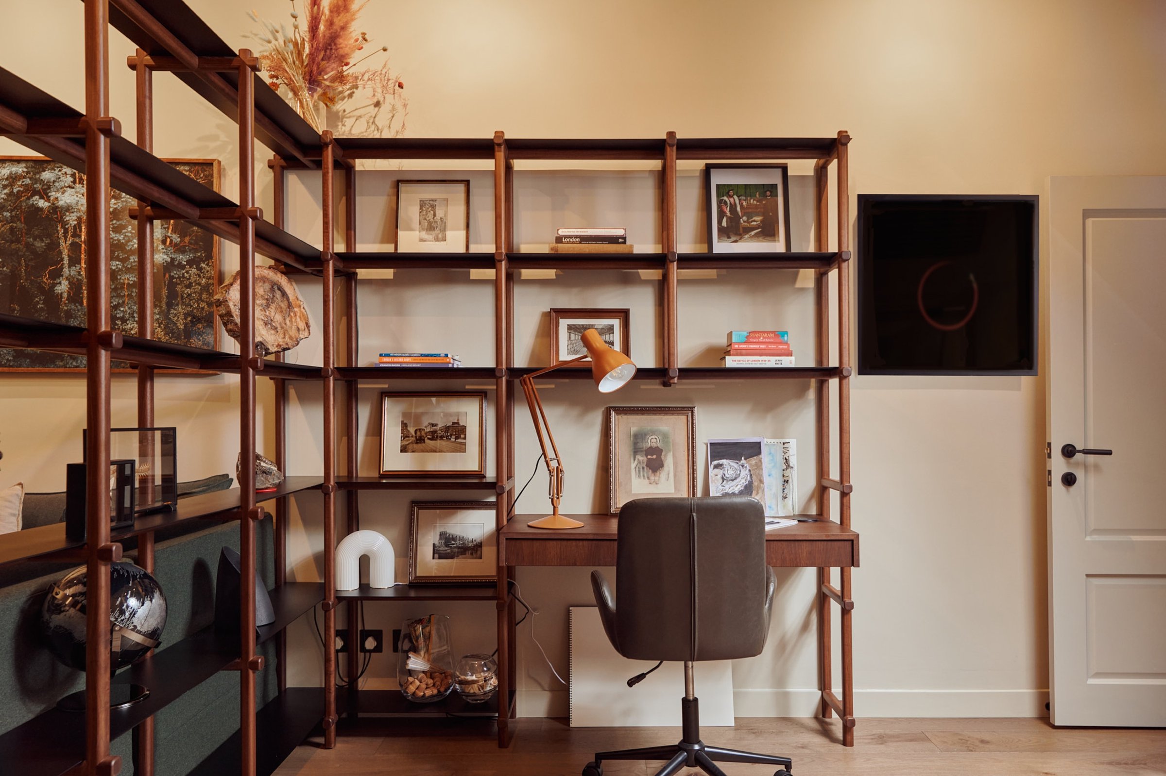 Study with a bolted timber shelving system across the wall, a desk built into it under an orange anglepoise, and framed pictures on the shelves.