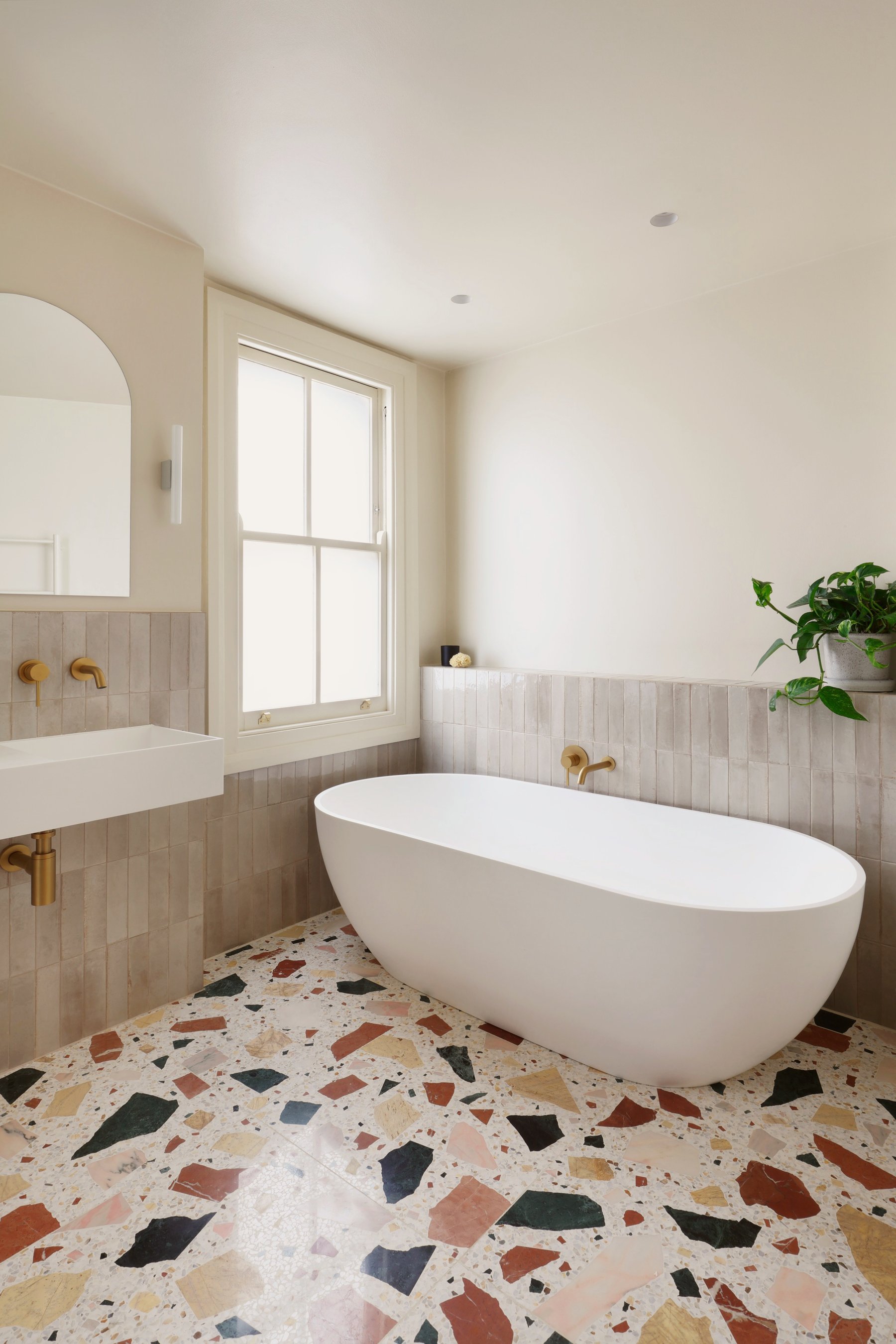 Freestanding white bath against half-height pale tiling with a brass floor-standing filler, a sash window to the left and terrazzo underfoot.