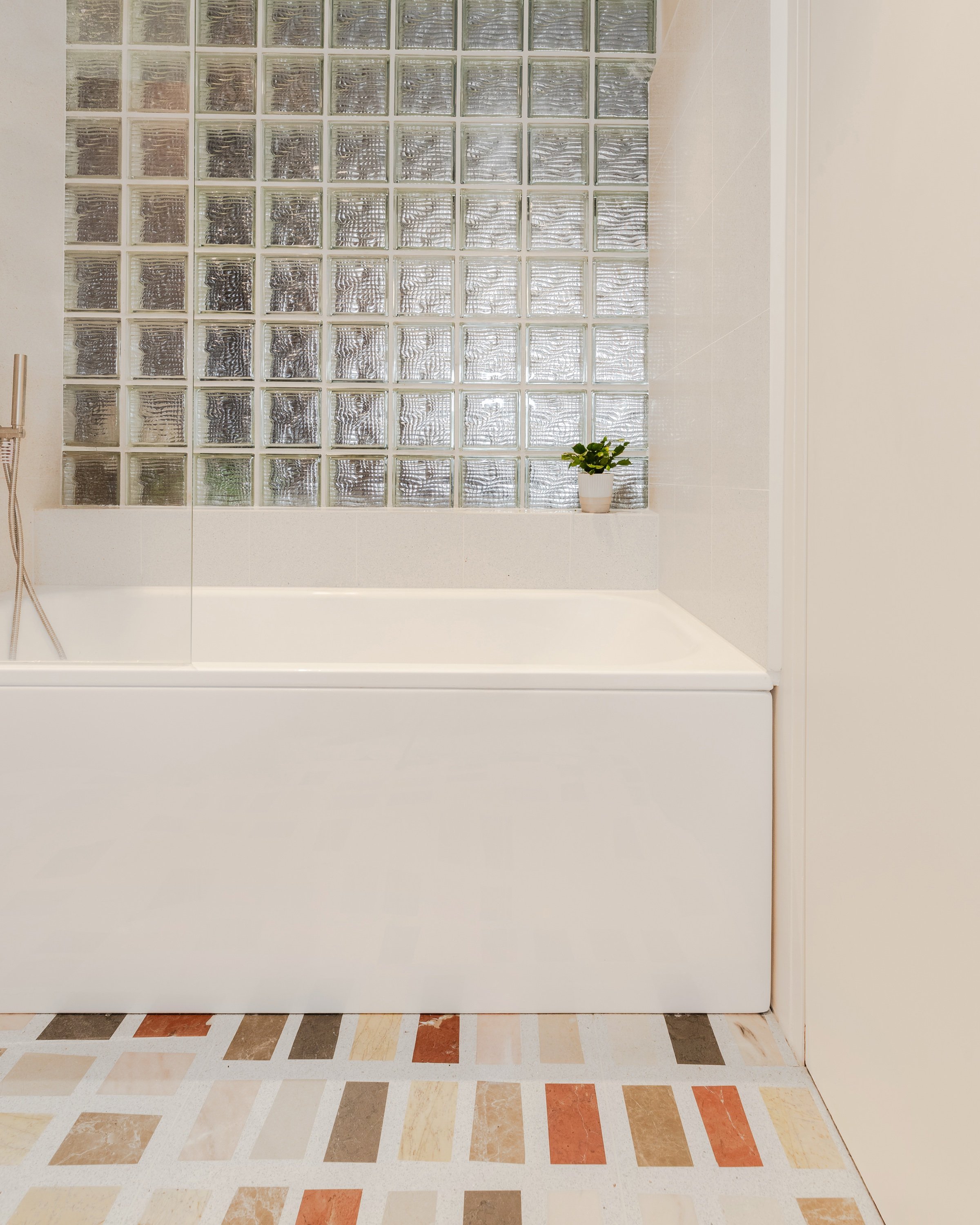 Bathroom with a white bath beneath an existing glass block window and a floor of rectangular marble tiles in orange, grey and cream.
