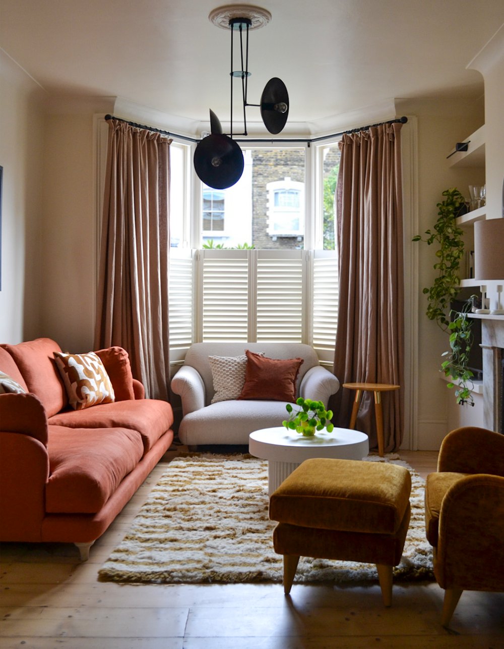 Living room in a bay-windowed Victorian front room, with plantation shutters, a terracotta sofa, dusty pink curtains and a sheepskin rug