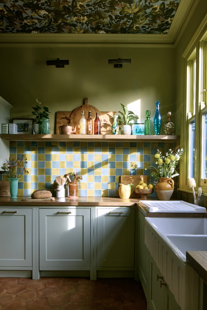 Kitchen with sage-green painted cabinets, a blue and yellow checked tile splashback, an oak shelf of jugs and bottles, and botanical wallpaper on the ceiling