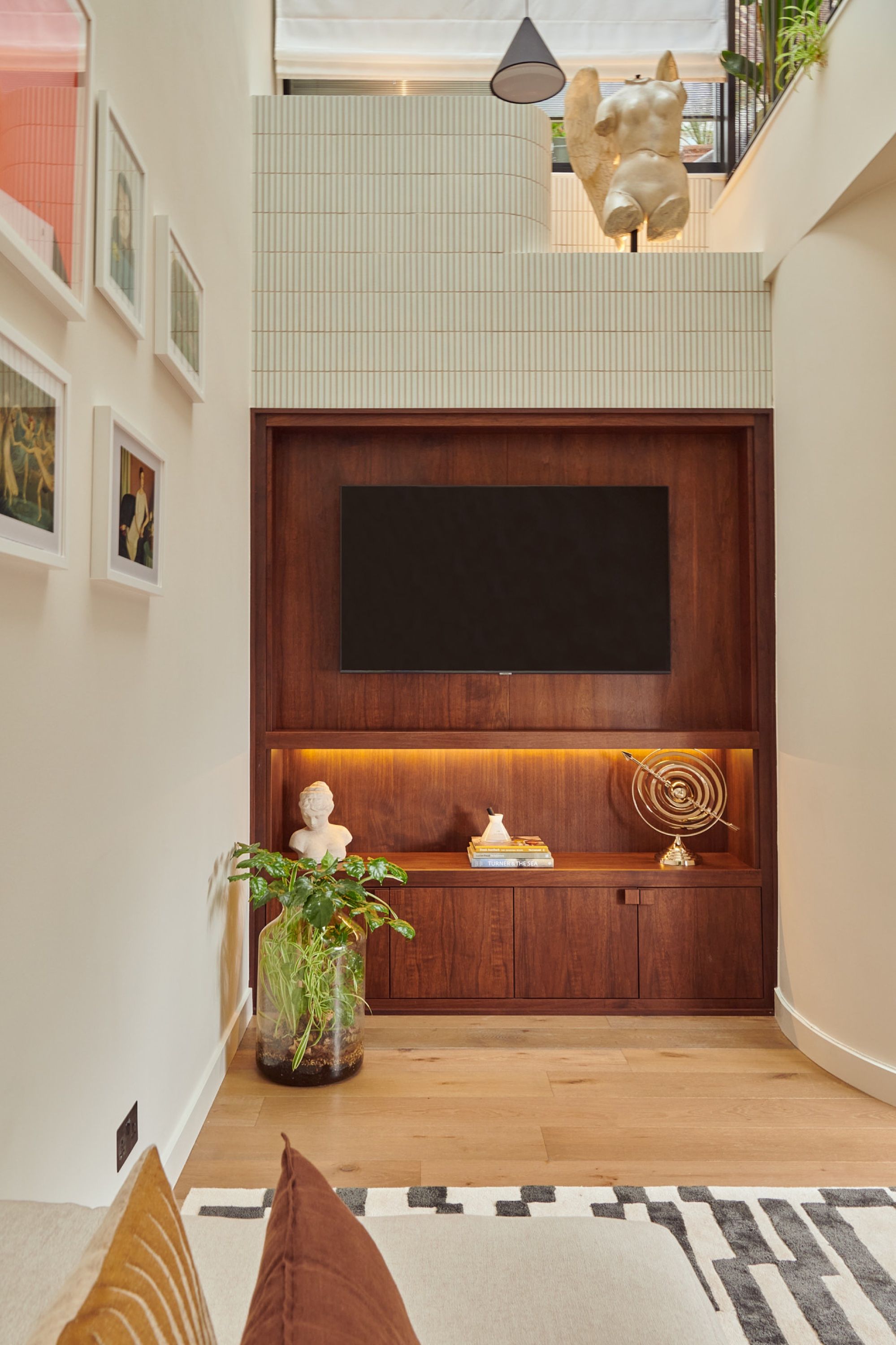 The walnut media recess straight on, its lit shelf holding a bust and a brass object, with a tiled upper level and sculpture on the landing above.