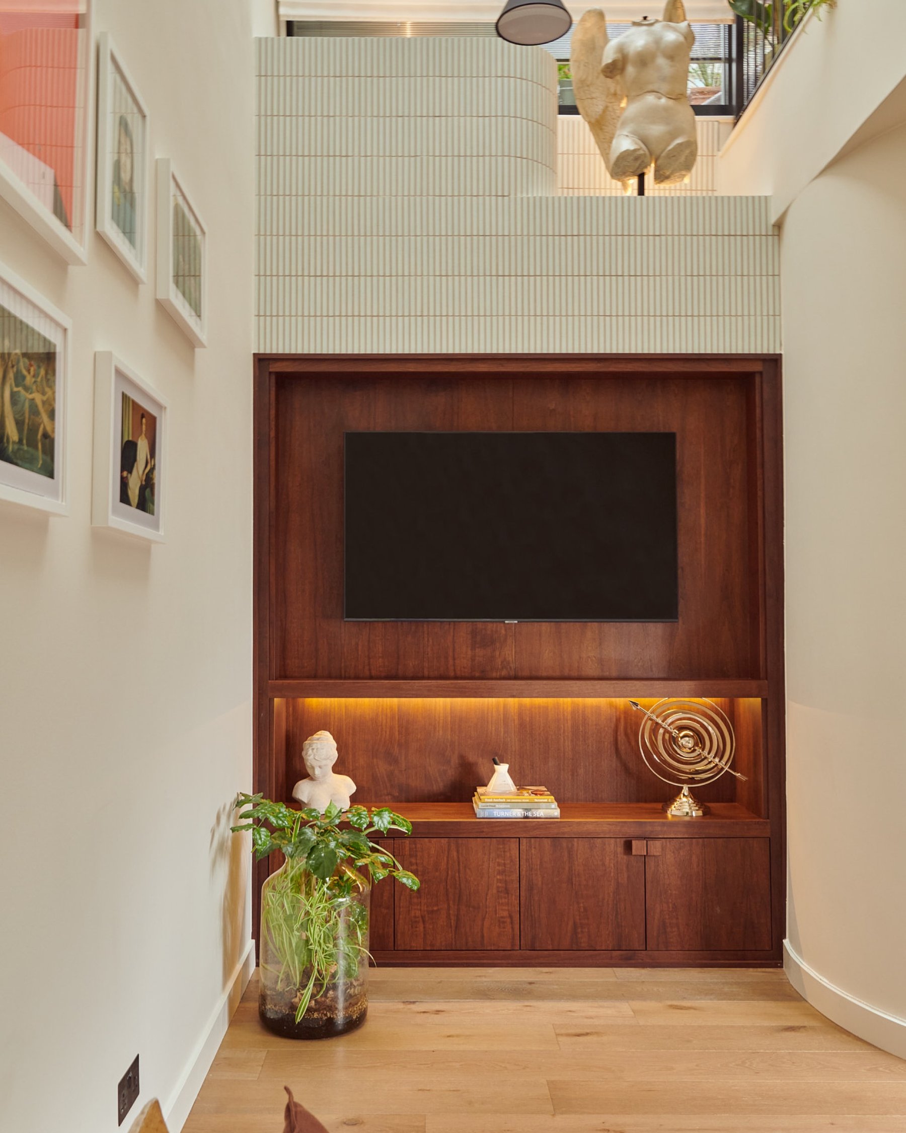 The walnut media recess straight on, its lit shelf holding a bust and a brass object, with a tiled upper level and sculpture on the landing above.