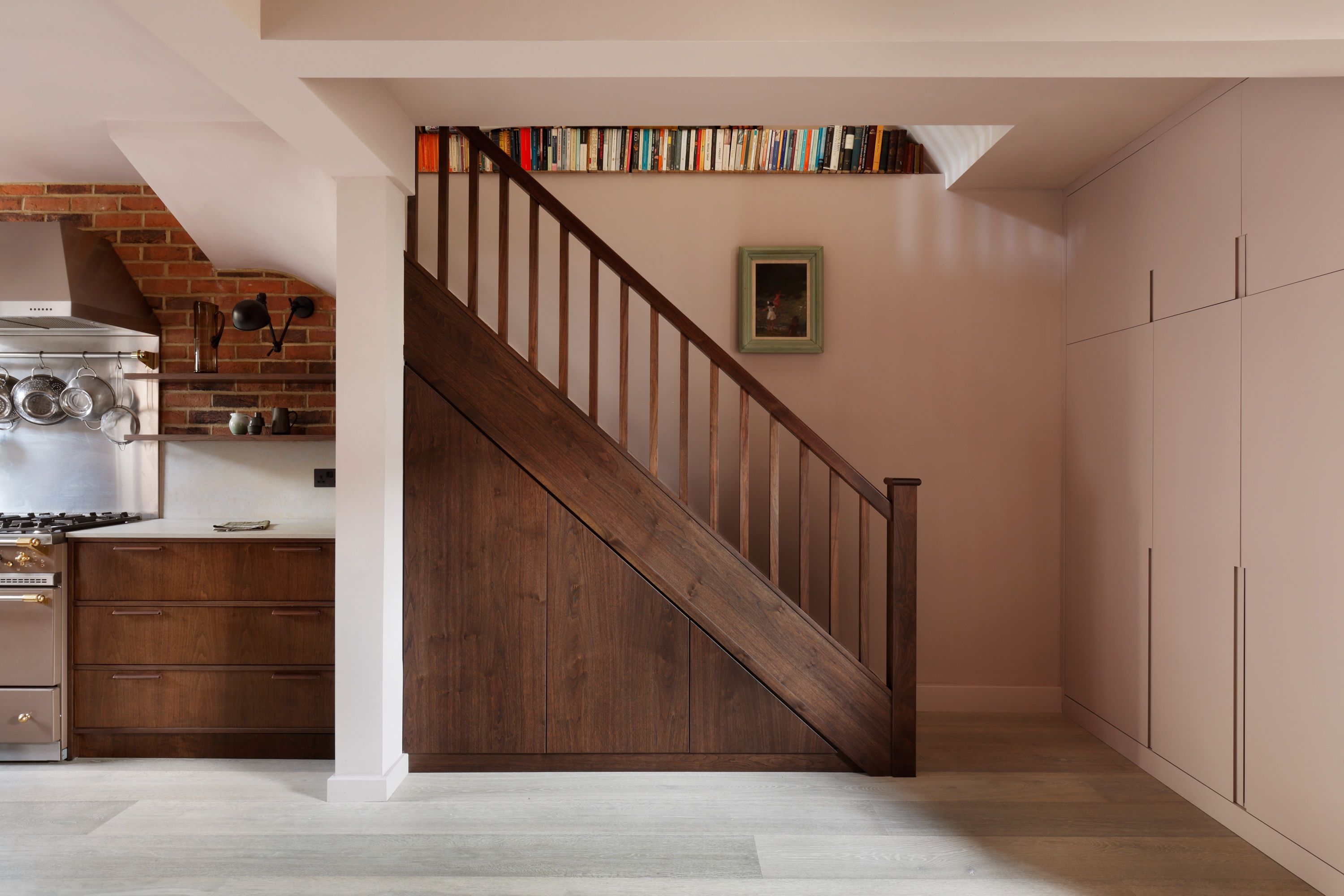 Dark stained timber staircase with a panelled under-stair face against a soft pink wall, books along the landing edge and the kitchen range left.