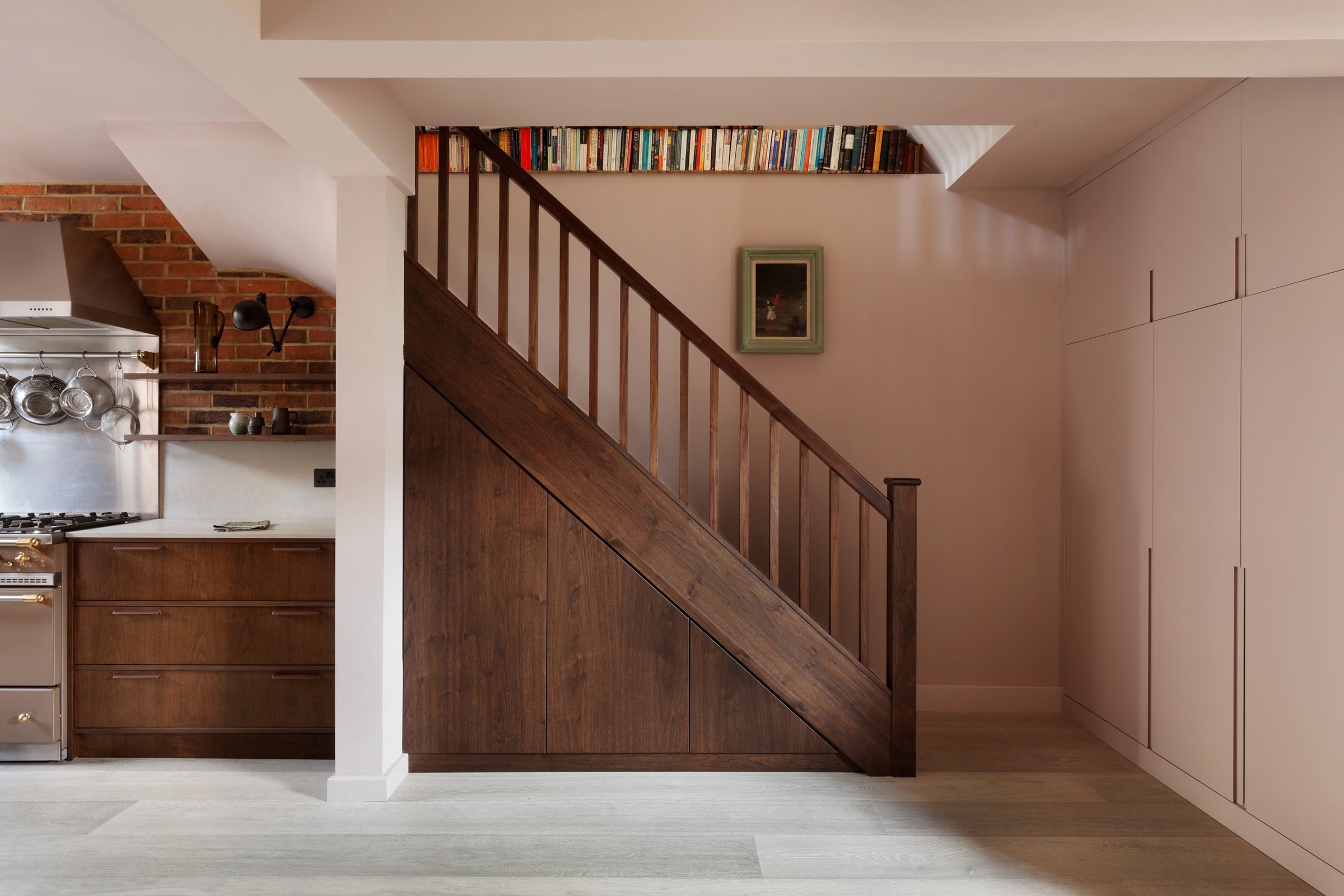 Dark stained timber staircase with a panelled under-stair face against a soft pink wall, books along the landing edge and the kitchen range left.
