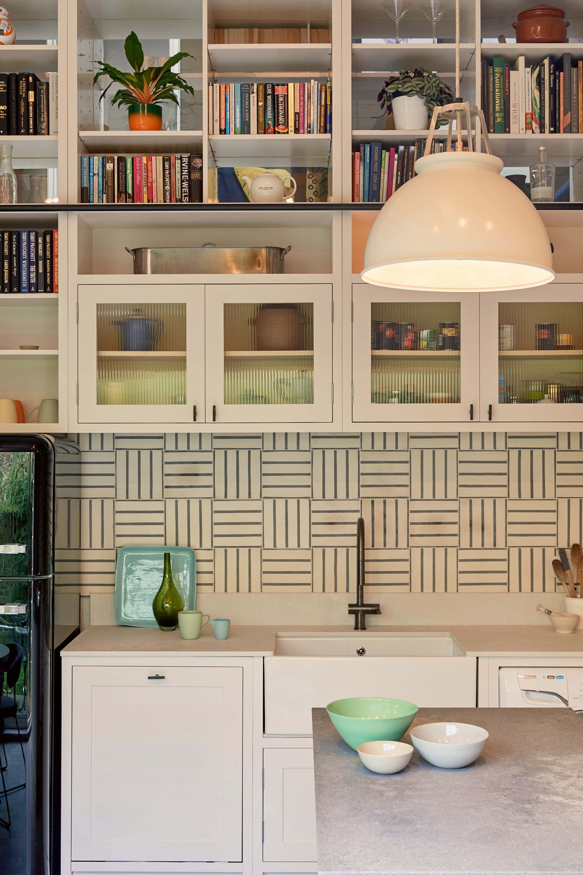 Kitchen sink wall with reeded glass cupboards, book-stacked shelves and a white pendant, the basketweave tiling lit beneath the cabinets.