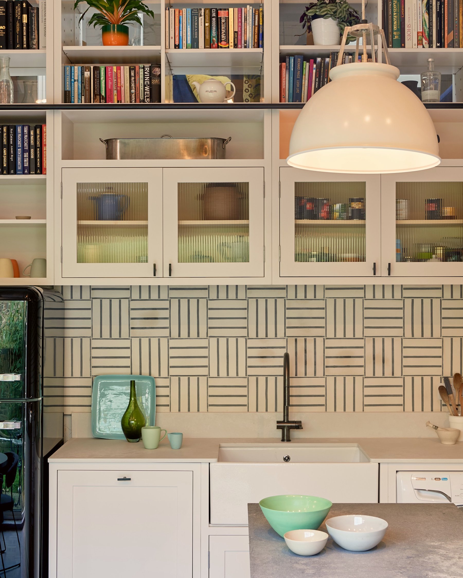 Kitchen sink wall with reeded glass cupboards, book-stacked shelves and a white pendant, the basketweave tiling lit beneath the cabinets.