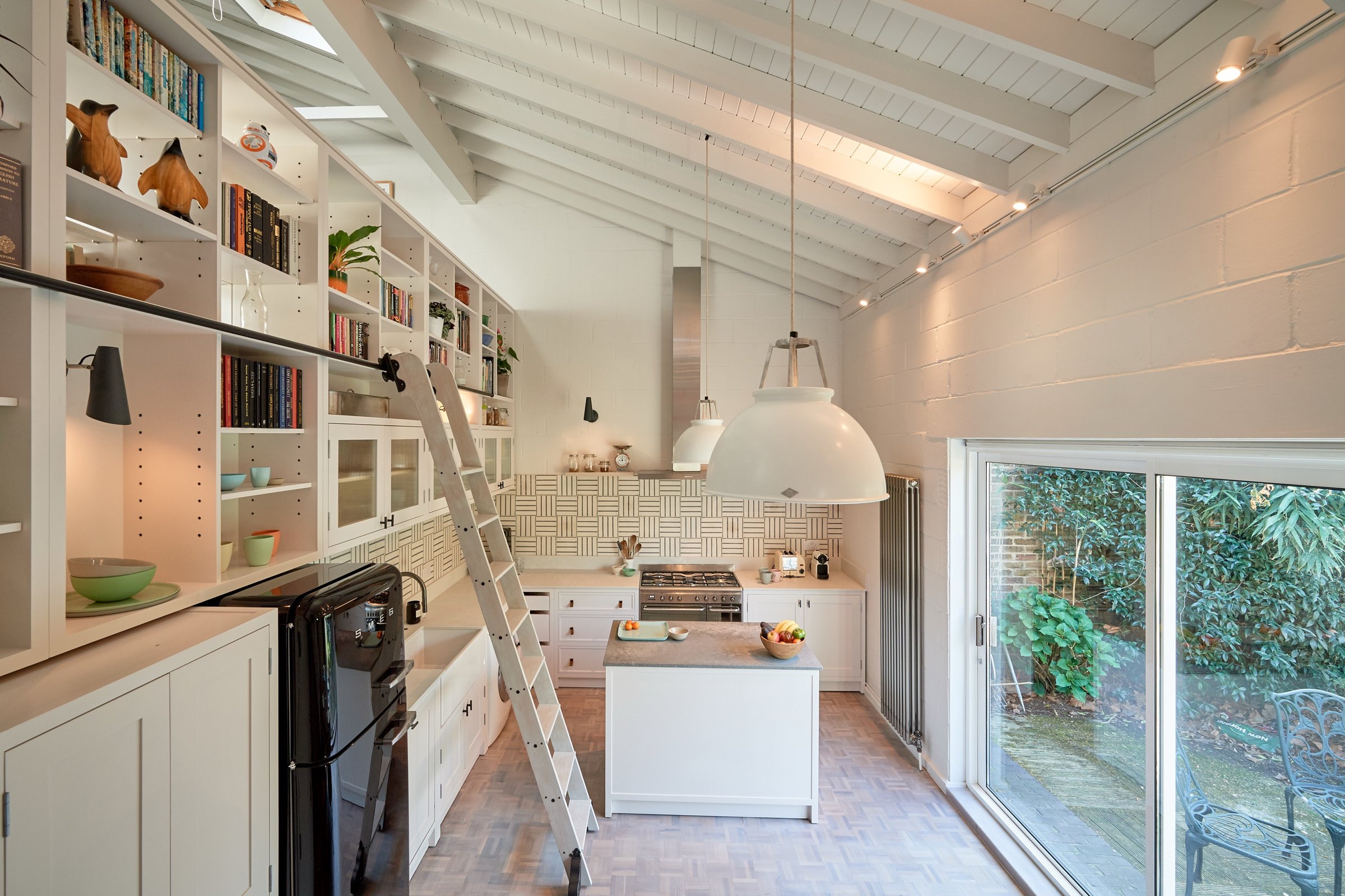 Long view of the kitchen, white joinery and library ladder down the left wall, island and range beyond, garden glazing at the right.