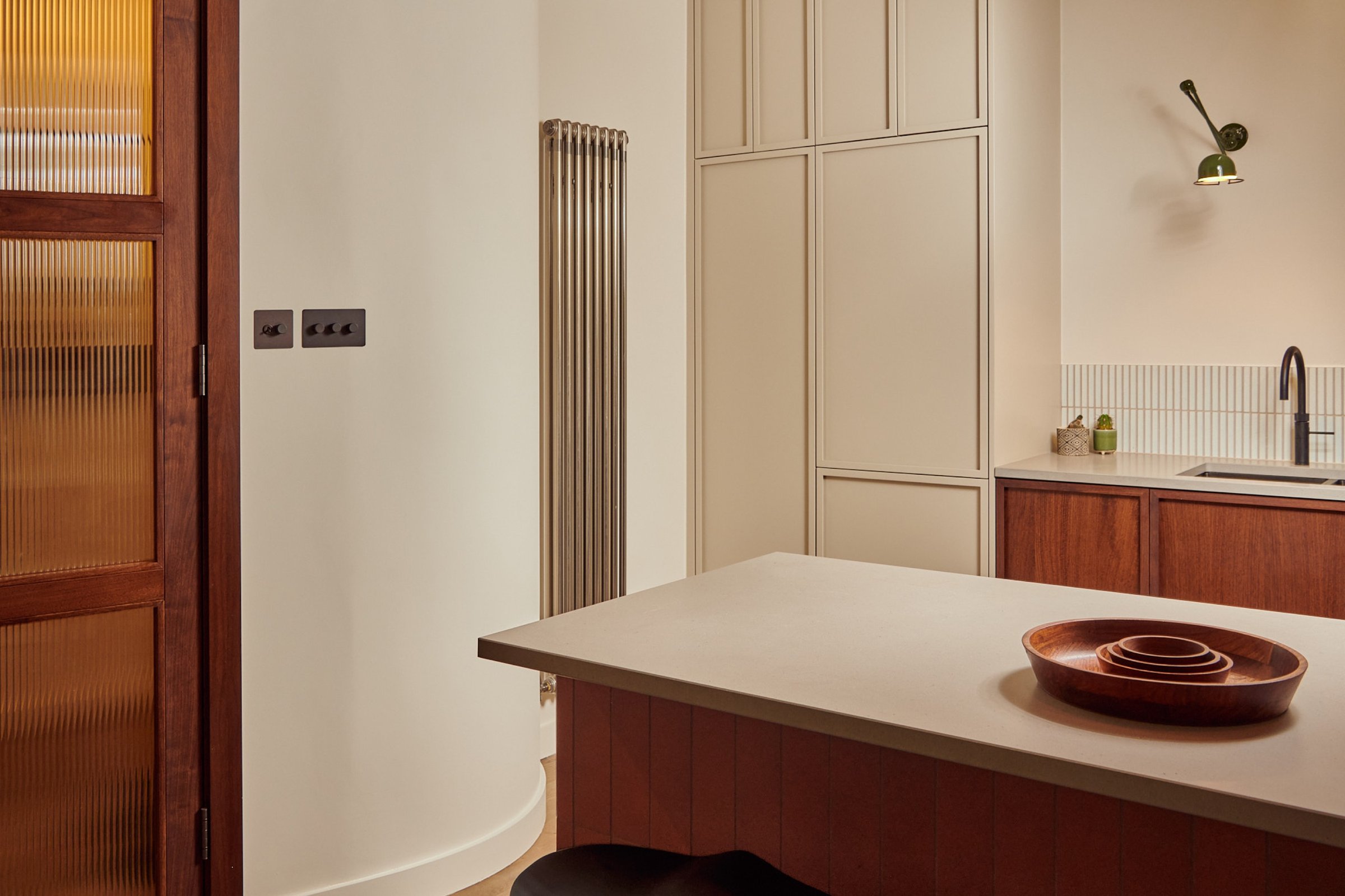 Kitchen past a reeded glass and timber door to a curved plastered wall and cream tall cupboards, with stacked wooden bowls on the island top.