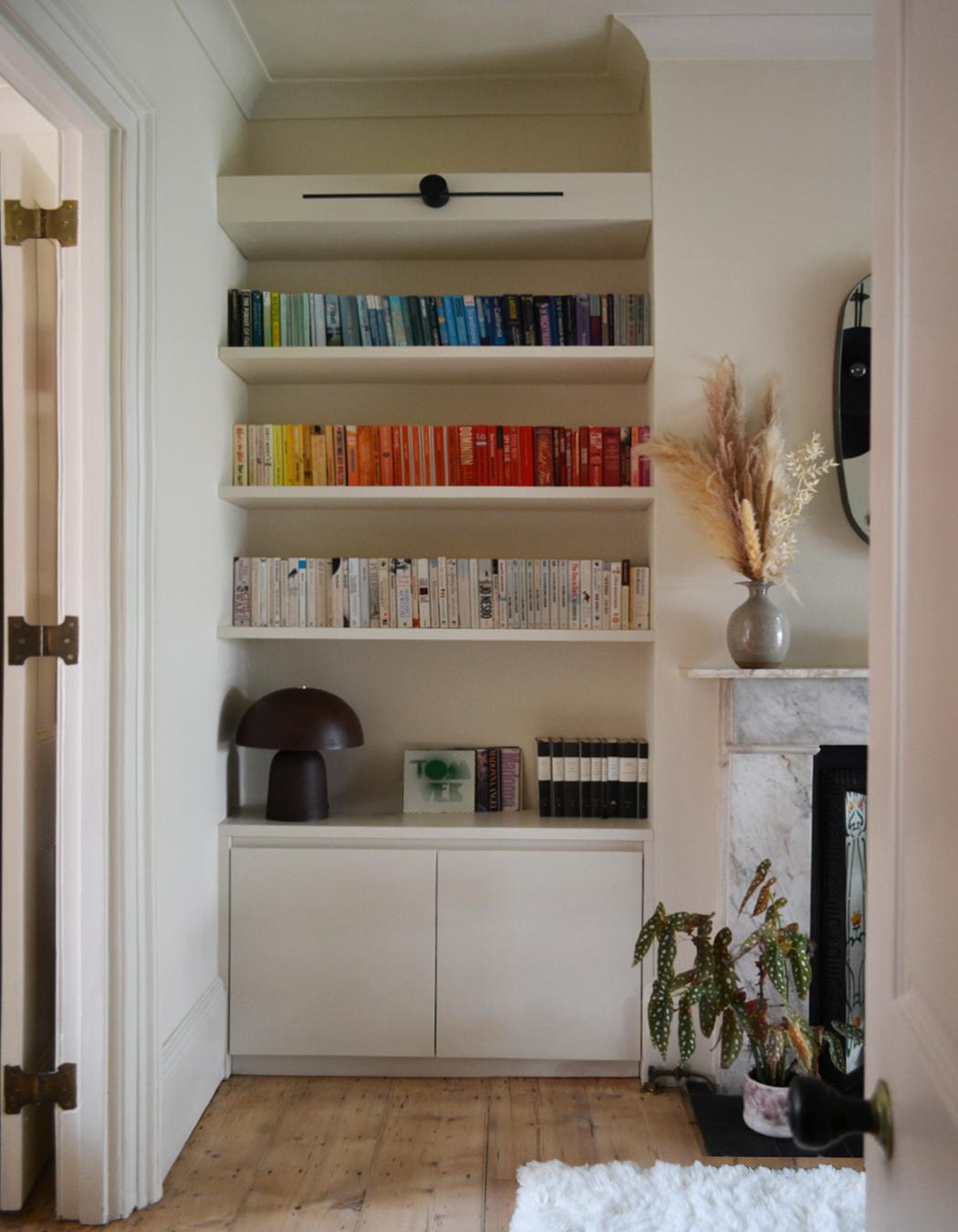 Alcove shelving filled with books arranged by colour, above fitted cupboards beside a marble fireplace