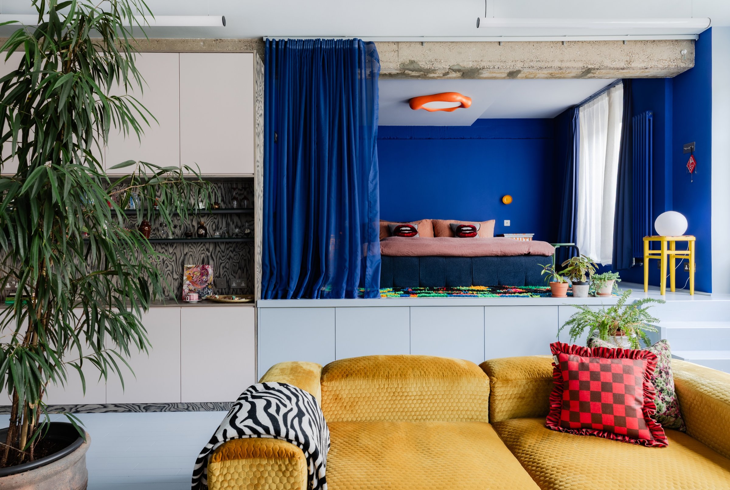 Wide view of the living space: pink cabinetry at the left, mustard modular sofa in front, and the blue sleeping platform raised behind a velvet curtain.