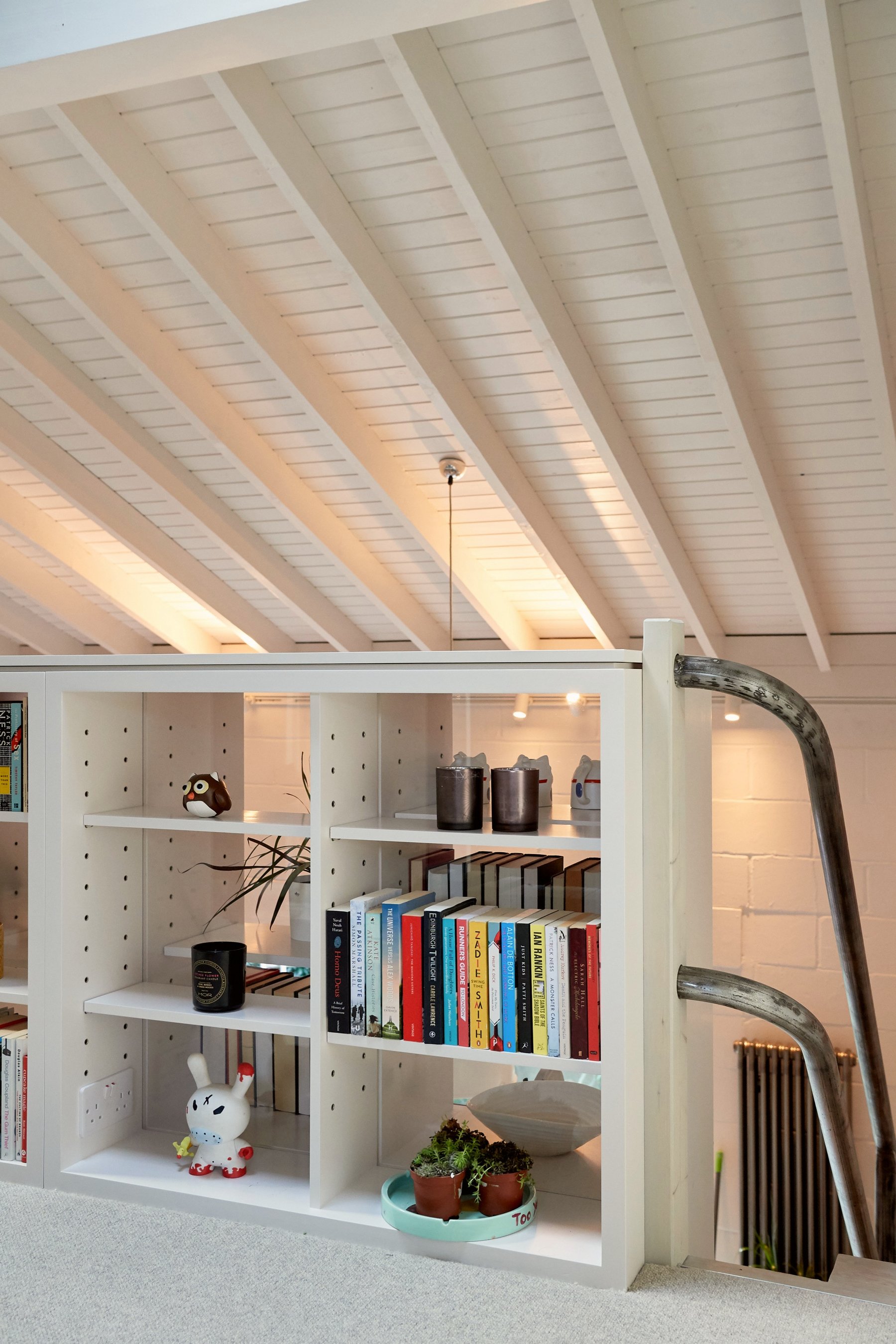 Mezzanine bookshelves built as a balustrade under a white-painted pitched and boarded roof, with an old iron handrail curving past.