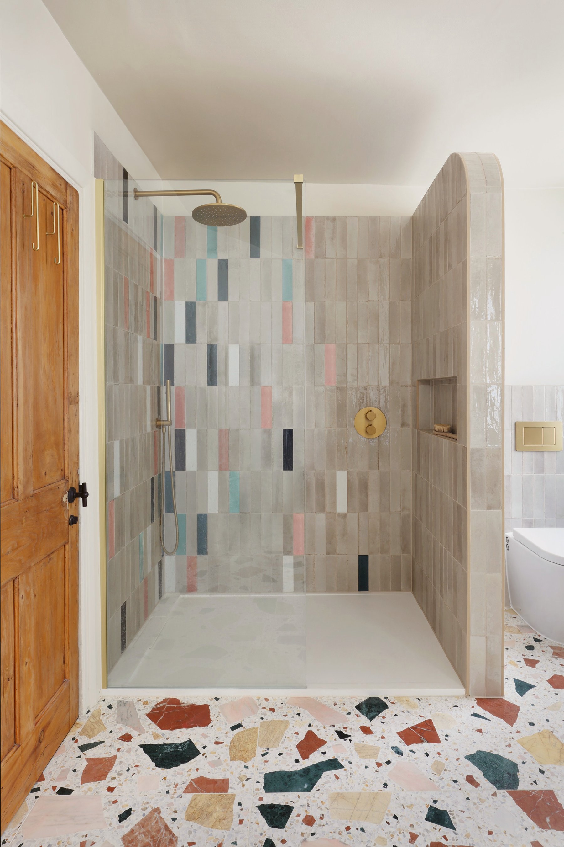 Walk-in shower tiled in narrow pale tiles scattered with pink, teal and navy, with a brass rainfall head, over a large-chip terrazzo floor.