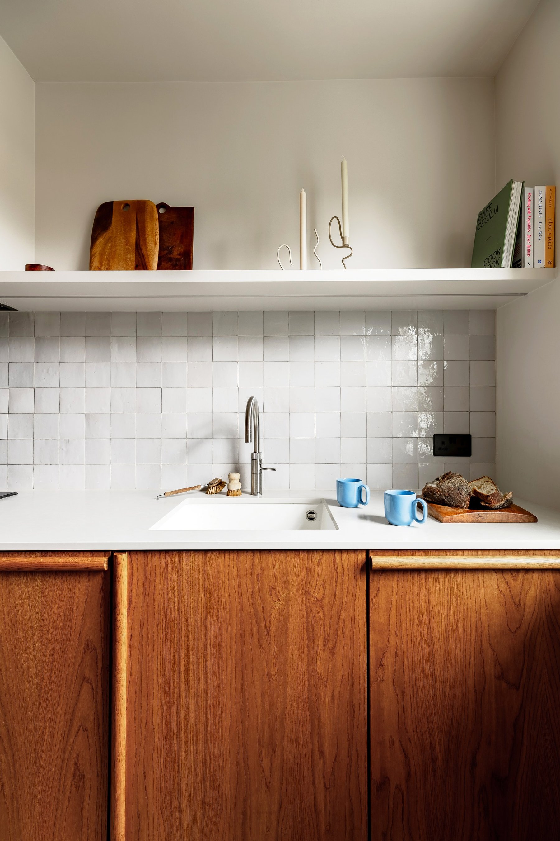 Kitchen sink run in wood cabinetry with rounded pulls, a white square-tiled splashback and an open shelf above.