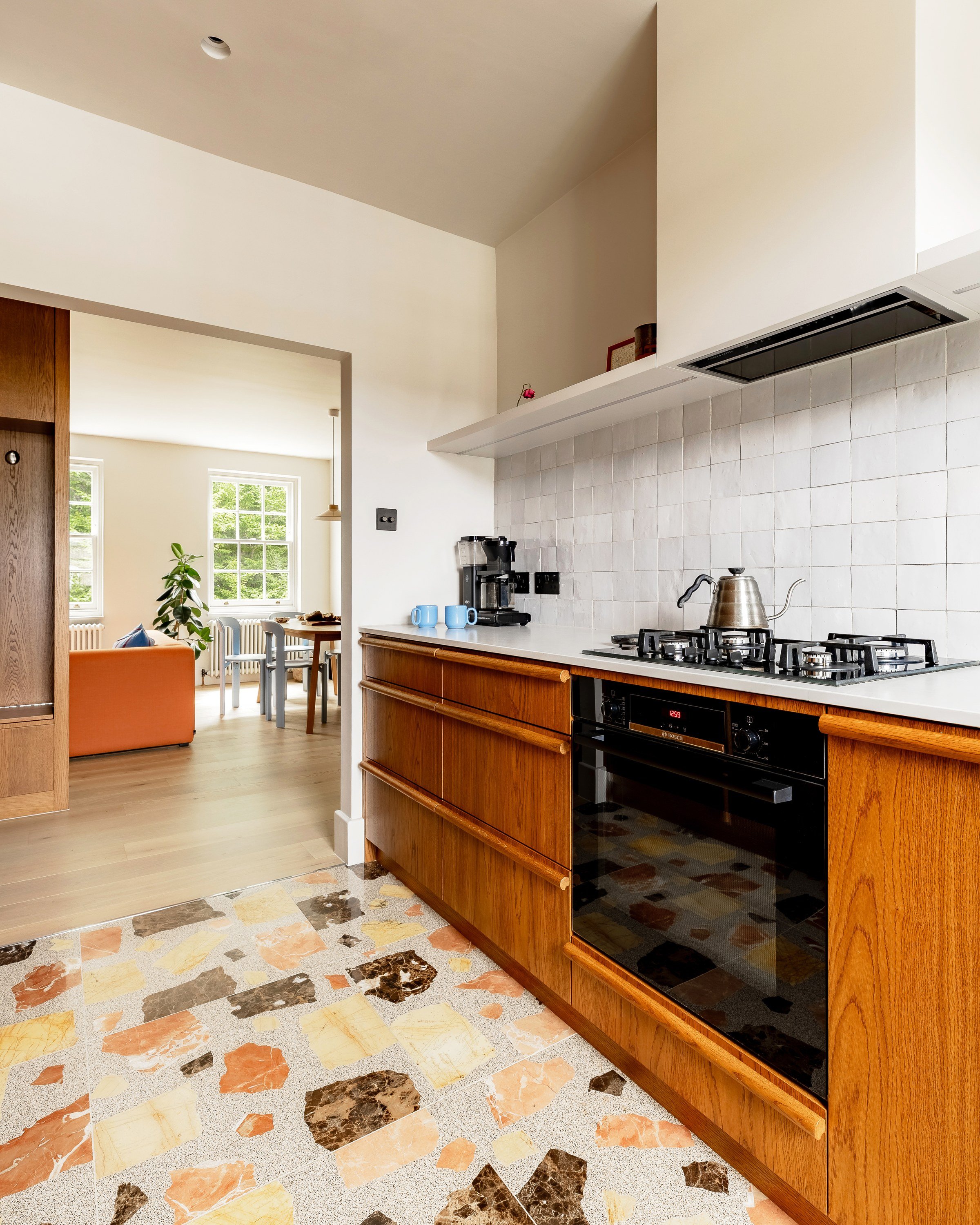Kitchen looking through to the living room: wooden units and a black oven on a terrazzo floor that meets pale boards at the doorway.