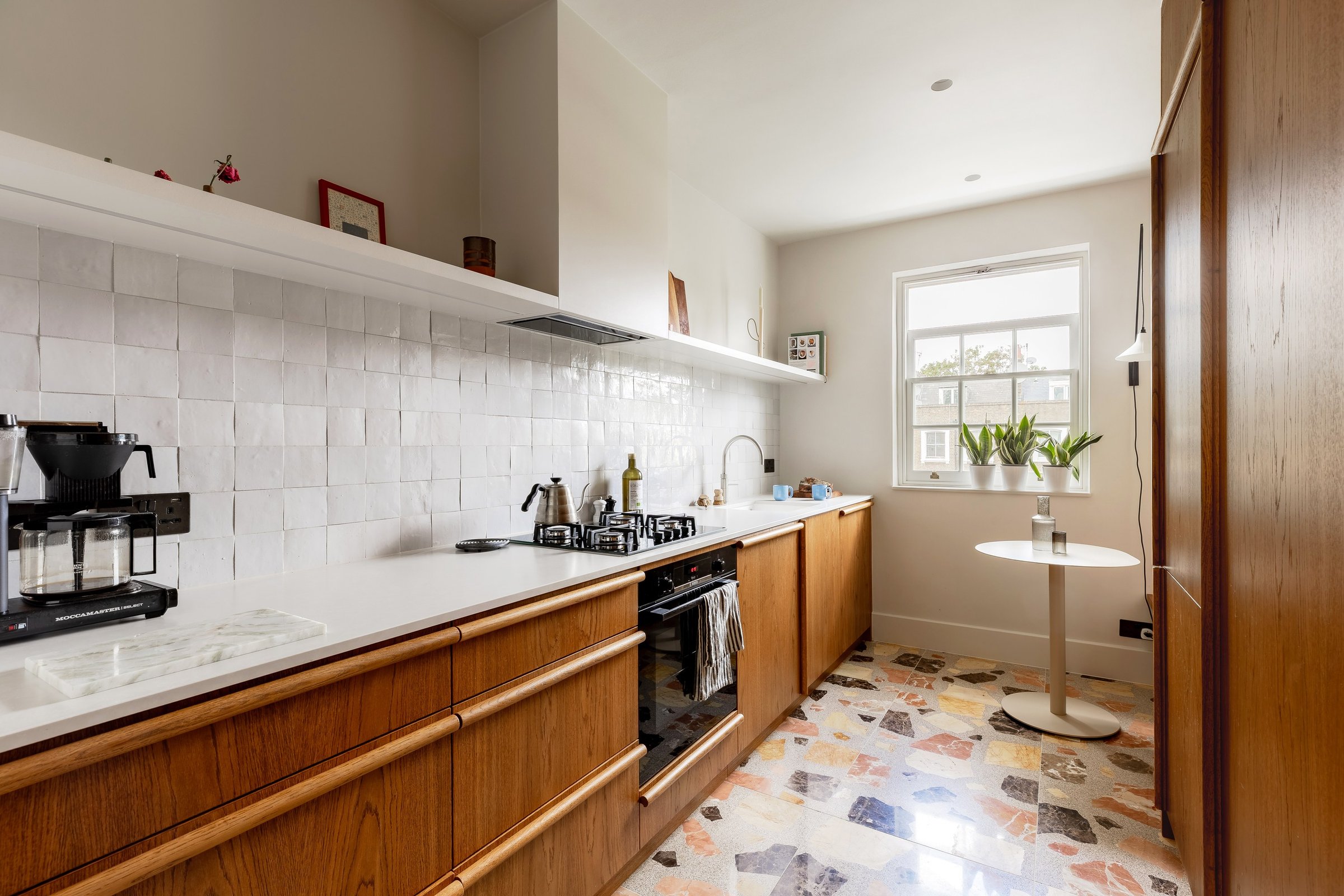 The galley kitchen down its length: wooden base units under a white worktop and tiled splashback, ending at a sash window with a small round table.