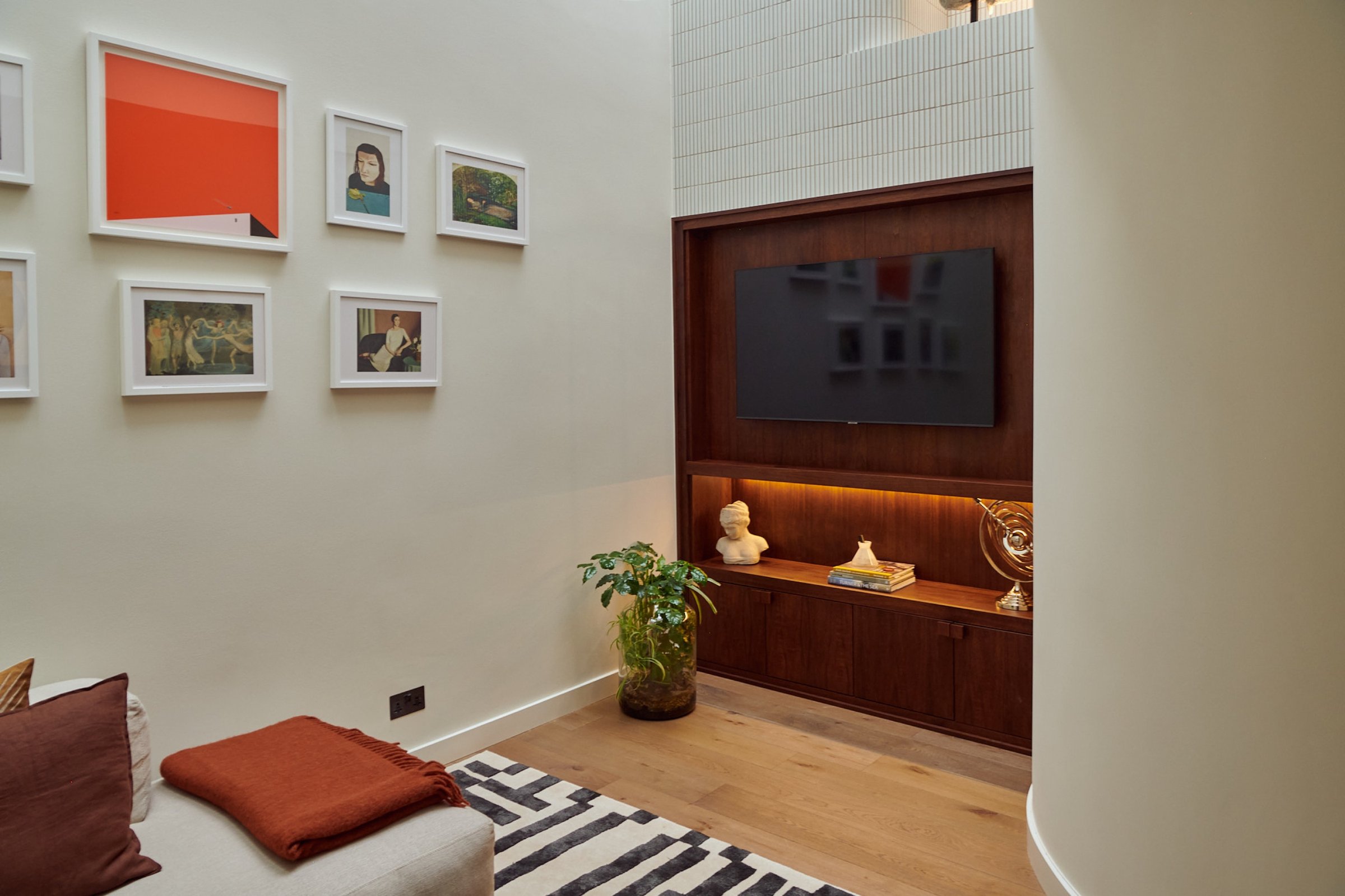 Sitting room corner with a walnut-lined media recess, a wall-mounted television above a lit shelf, and framed pictures on the adjacent wall.