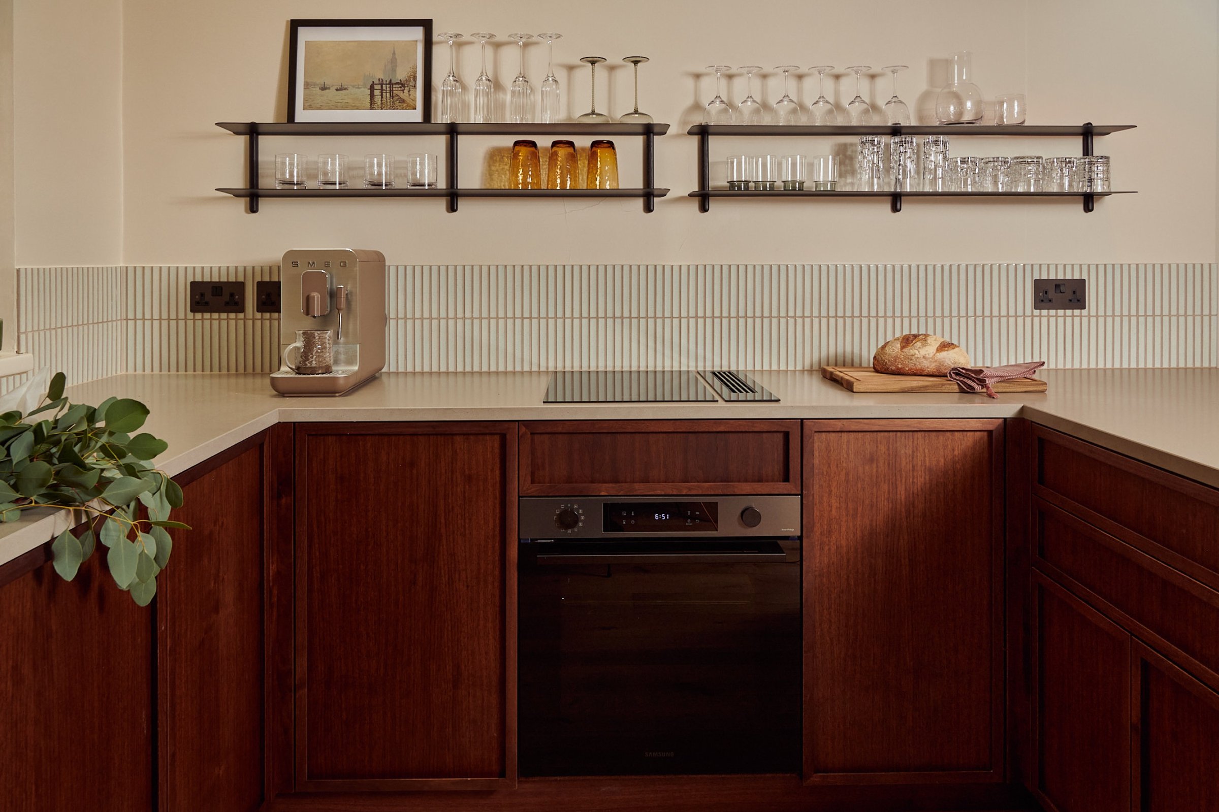The walnut run straight on: oven below the hob, fluted white splashback, and two long black shelves holding glasses and a carafe.