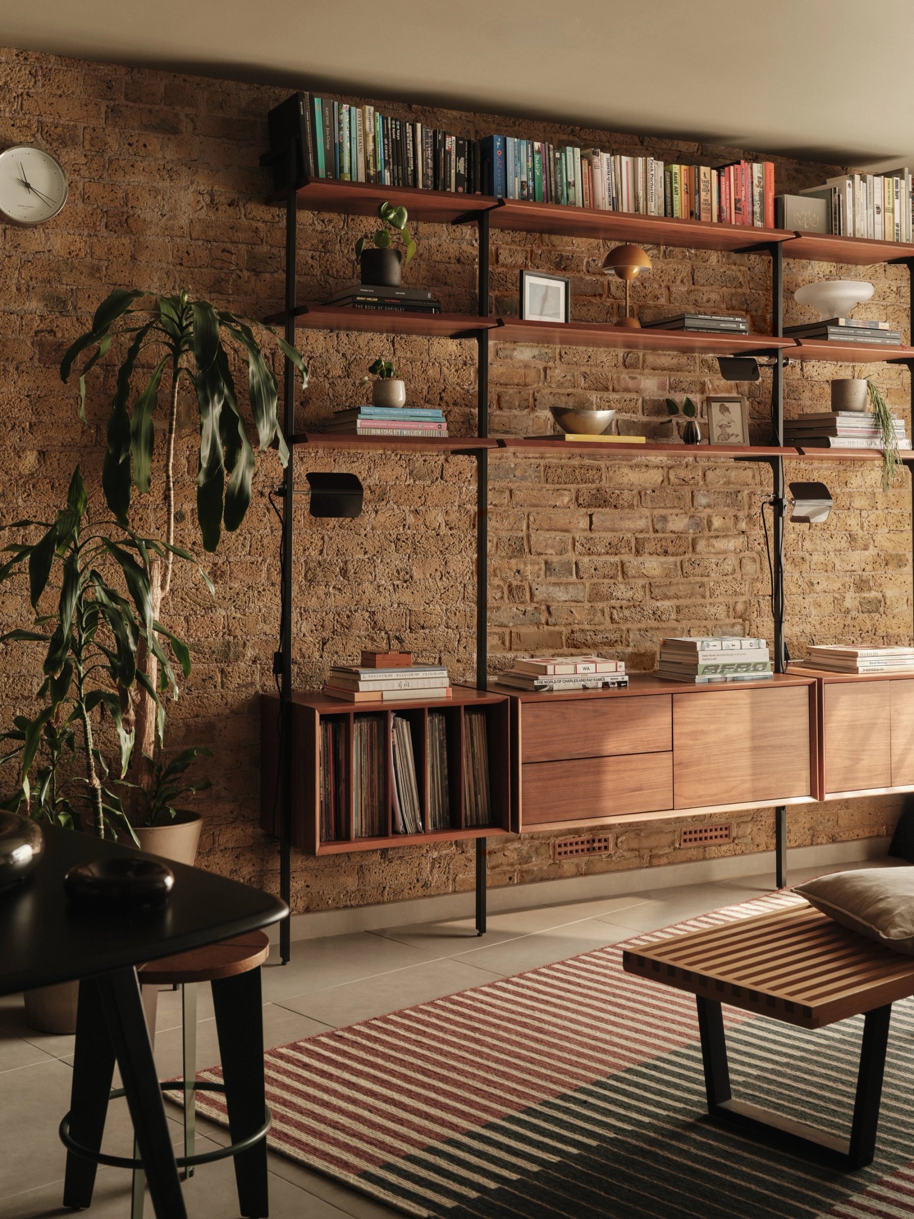Sitting room with a wall-hung teak shelving system on black uprights against exposed brick, books and records above a run of teak cabinets.