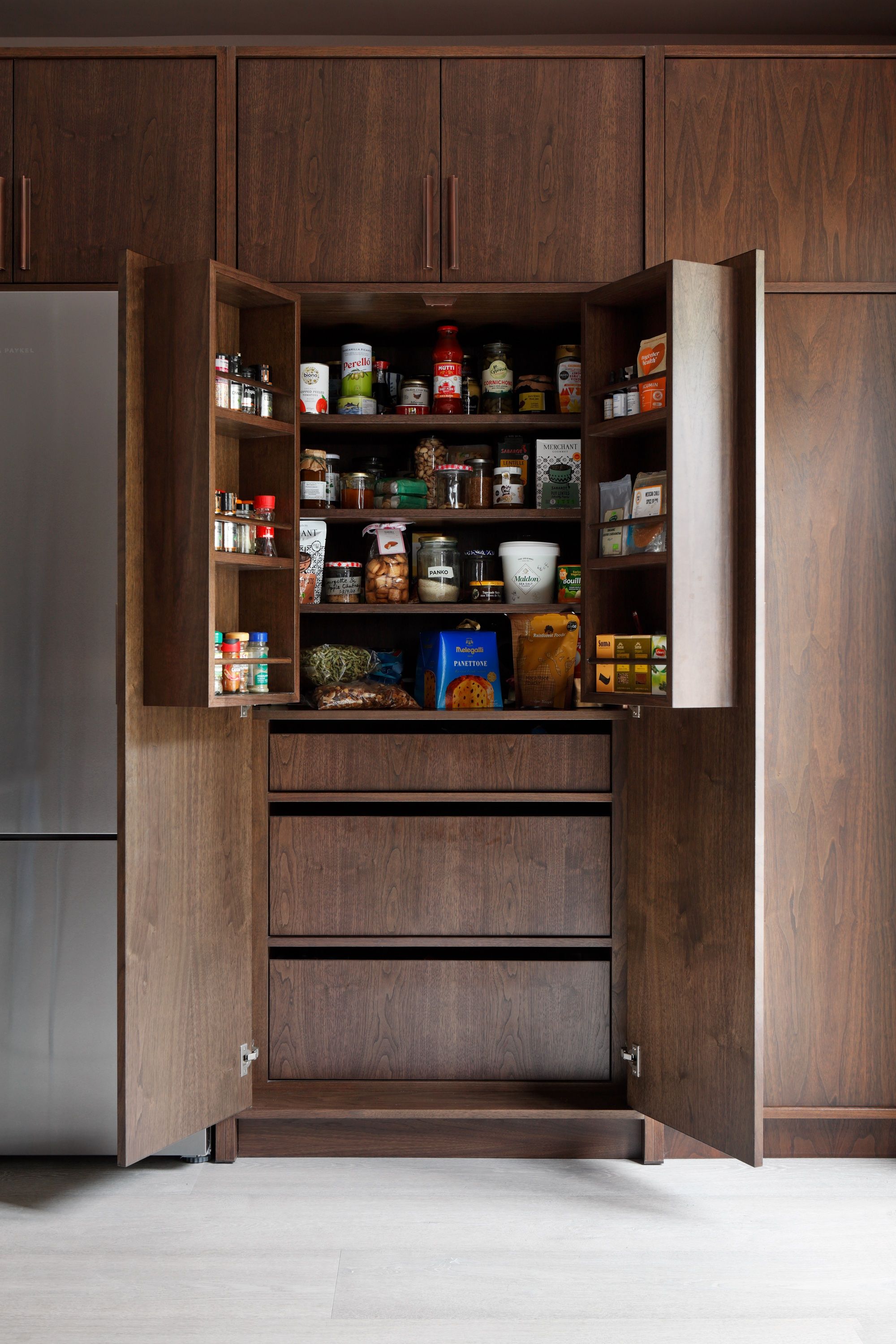 Tall dark timber larder standing open, its door racks and shelves stocked with jars and packets, drawers below and a fridge alongside.