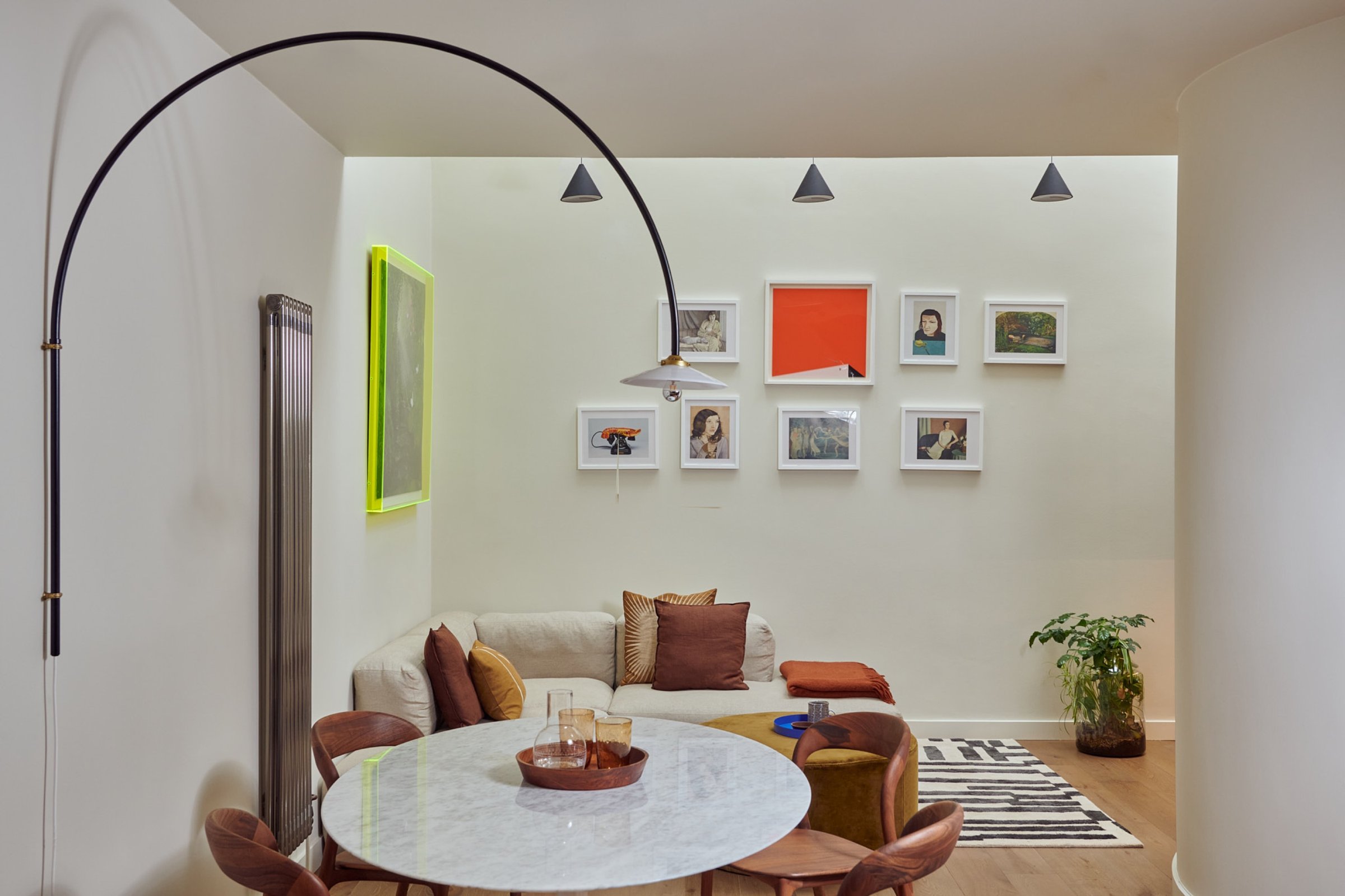 Open-plan sitting and dining area with a round marble table, a corner sofa beneath a salon hang of small frames, and an arc floor lamp overhead.