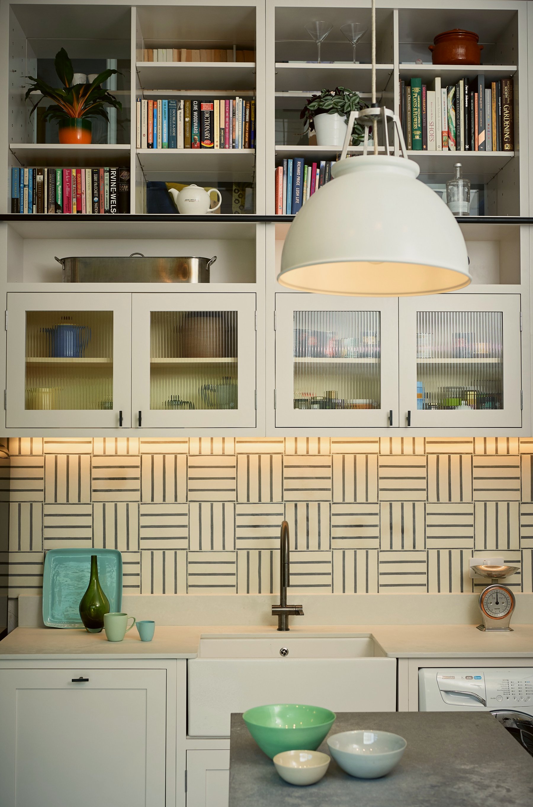 Kitchen sink wall straight on, butler sink and gunmetal tap below lit basketweave tiling, with reeded glass cupboards and books above.