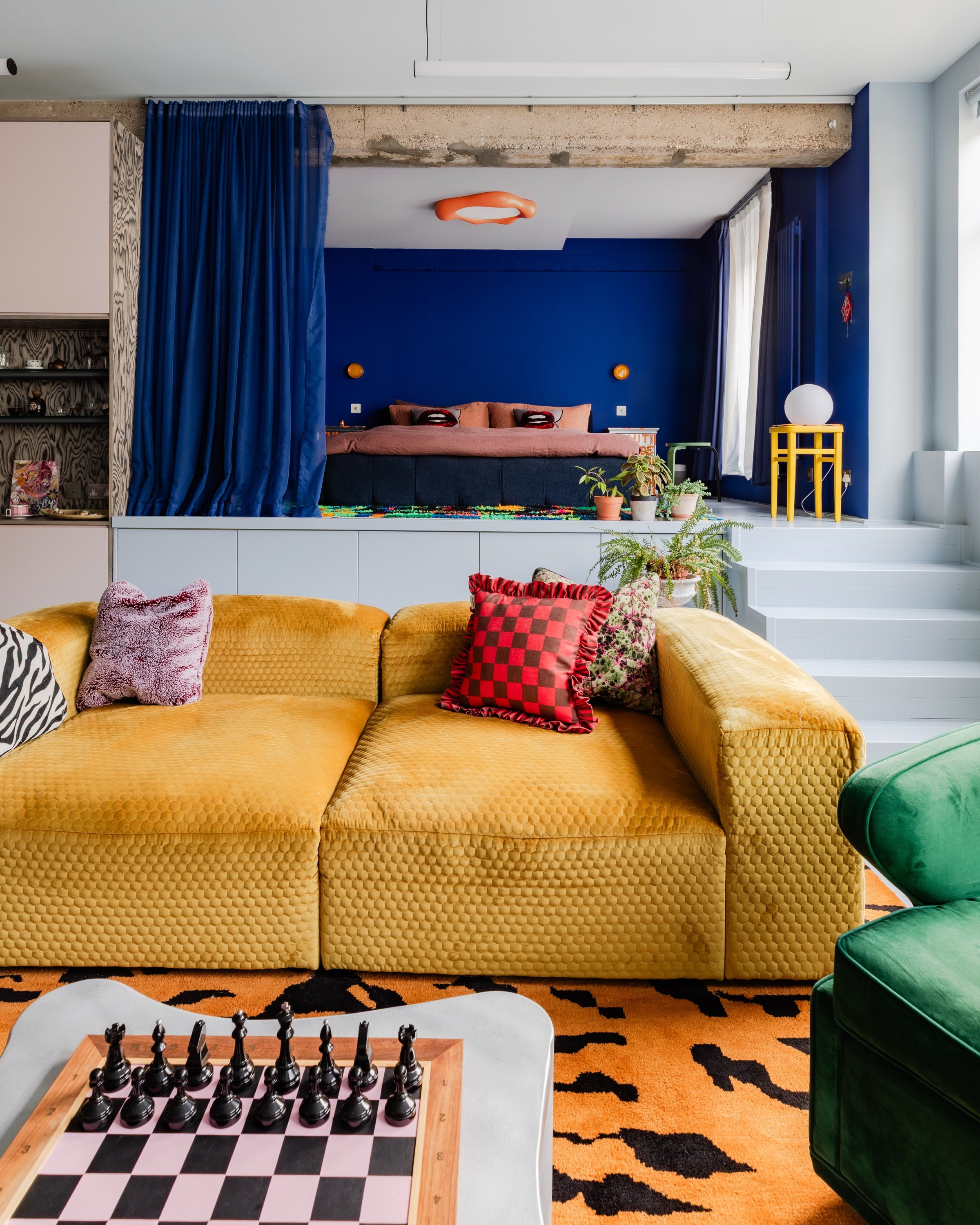 Living area looking across a mustard velvet modular sofa to a raised sleeping platform behind a blue curtain, under an exposed concrete beam.