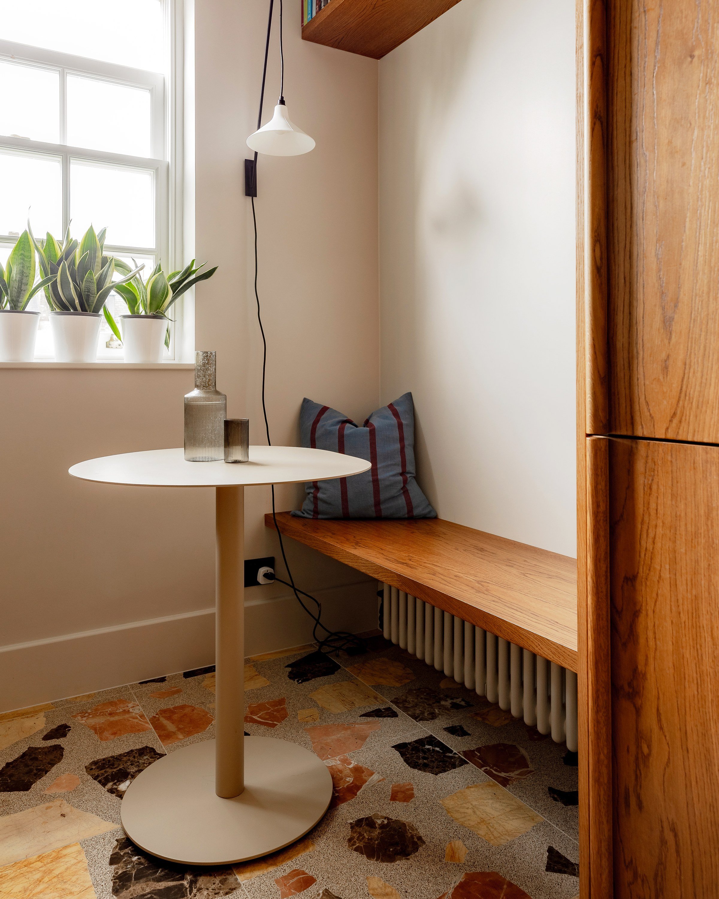 Window seat in wood with a striped cushion, a round white table and plants on the sill at the end of the kitchen.