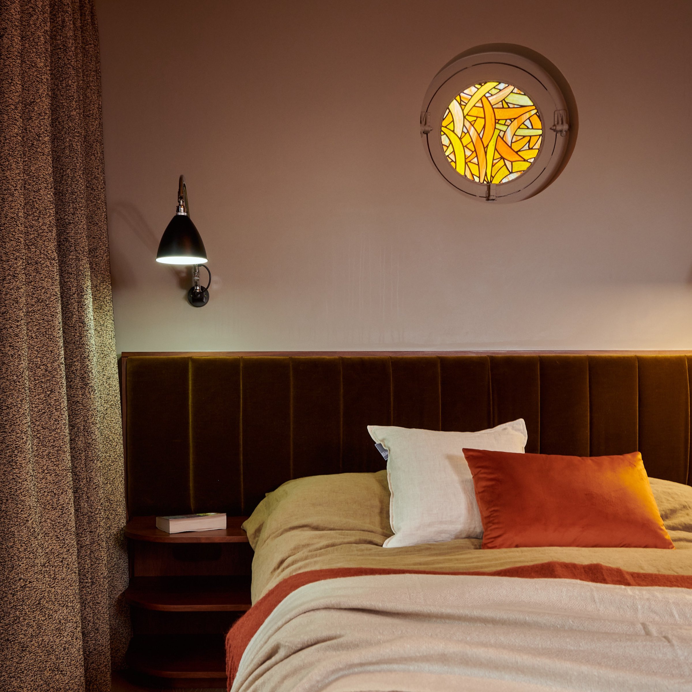 Bedhead detail: a fluted olive velvet headboard against a soft pink wall, with an amber stained glass porthole window above.