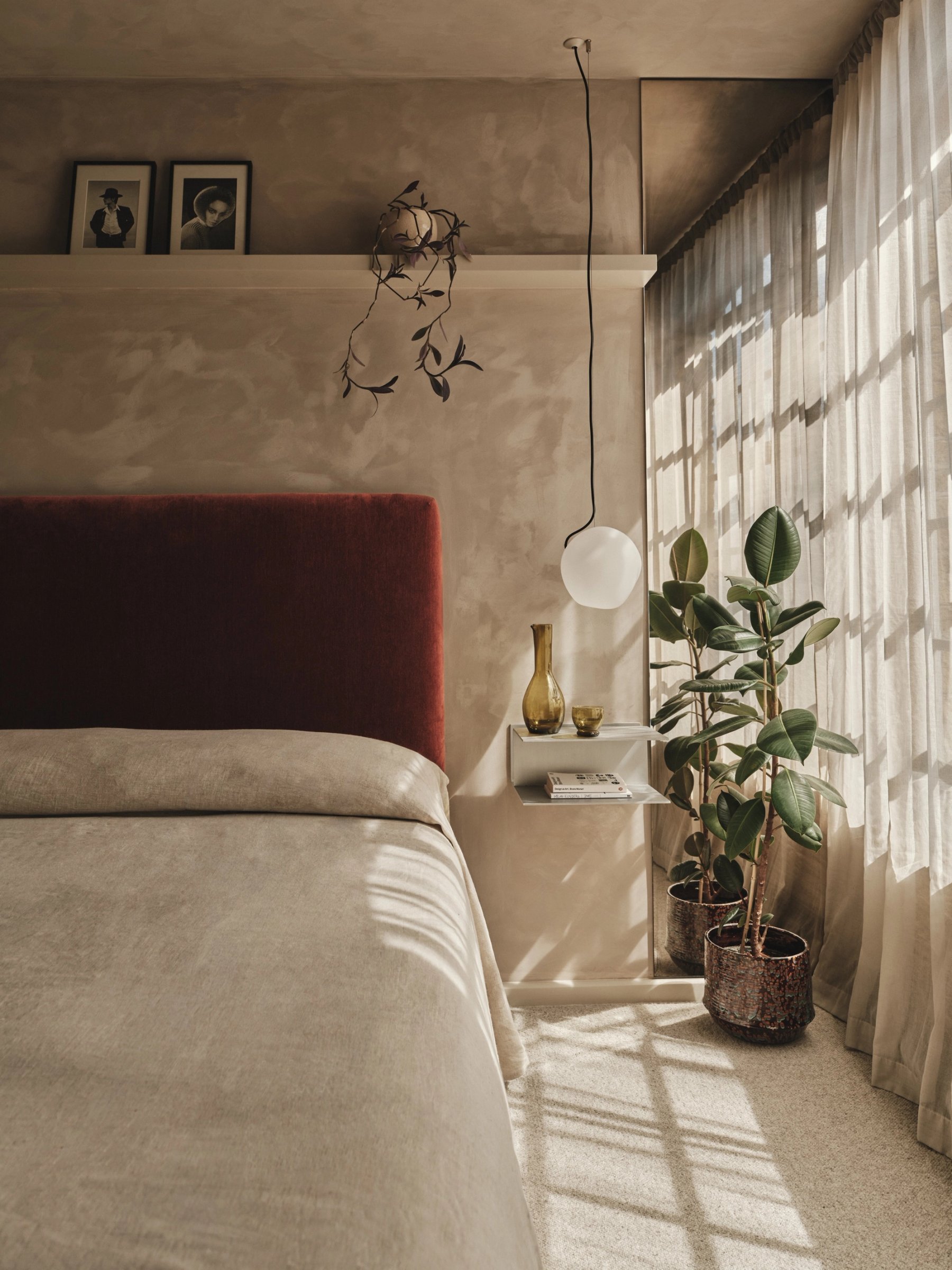 Bedroom corner with a rust velvet headboard against mottled polished plaster, a folded-metal bedside shelf and a globe pendant on a long flex.