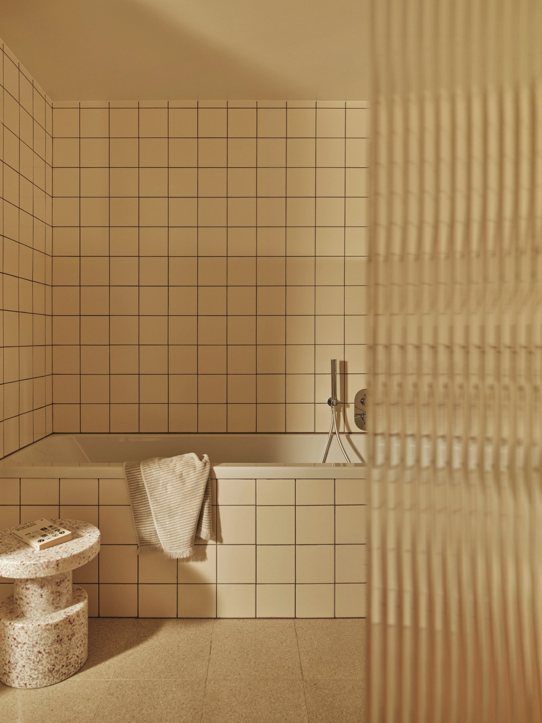 Bathroom tiled floor to ceiling in square cream tiles, a bath set into the tiling with a slim chrome filler and a reeded glass screen in front.