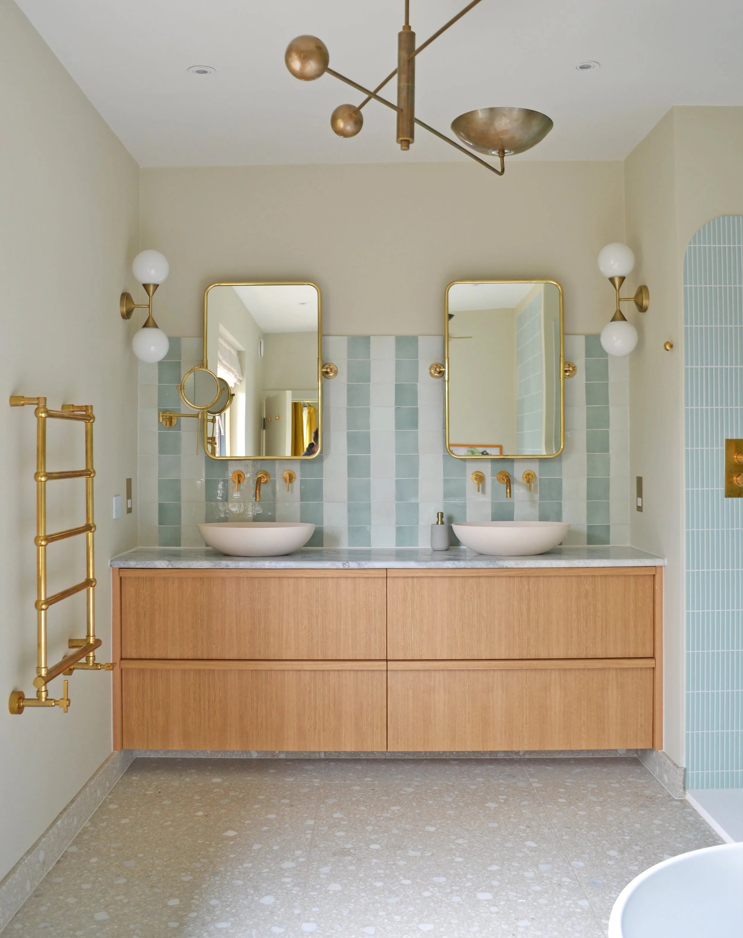 Bathroom with a double oak vanity, twin marble-topped basins and brass fittings, against pale blue vertical tiles on a terrazzo floor