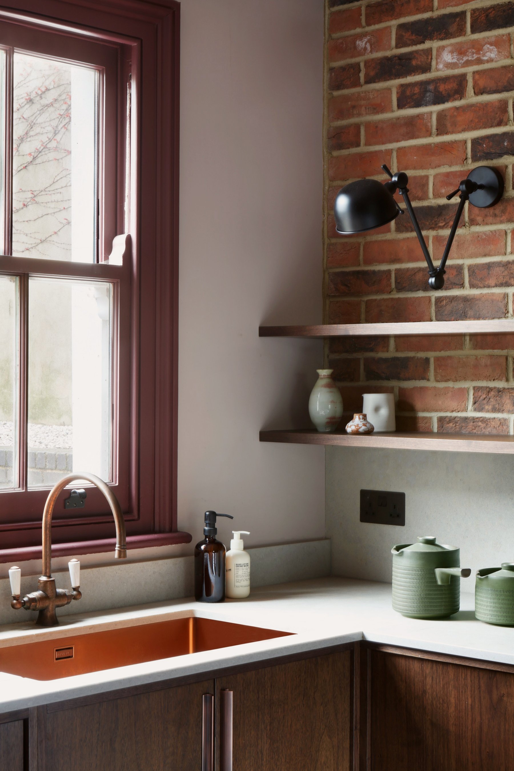Kitchen corner with a copper sink and tap below a red-framed sash window, timber shelves against exposed brick and a black wall light above.