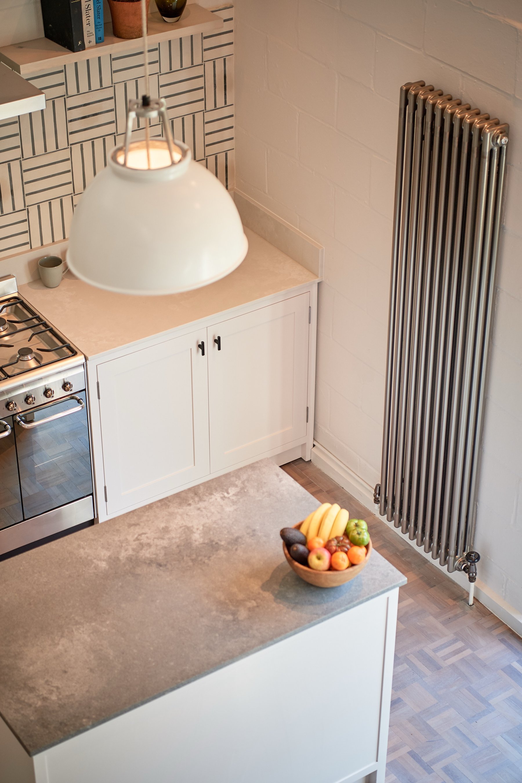 Kitchen detail looking down at the island's grey worktop with a bowl of fruit, the range cooker left and a slim column radiator against painted brick.