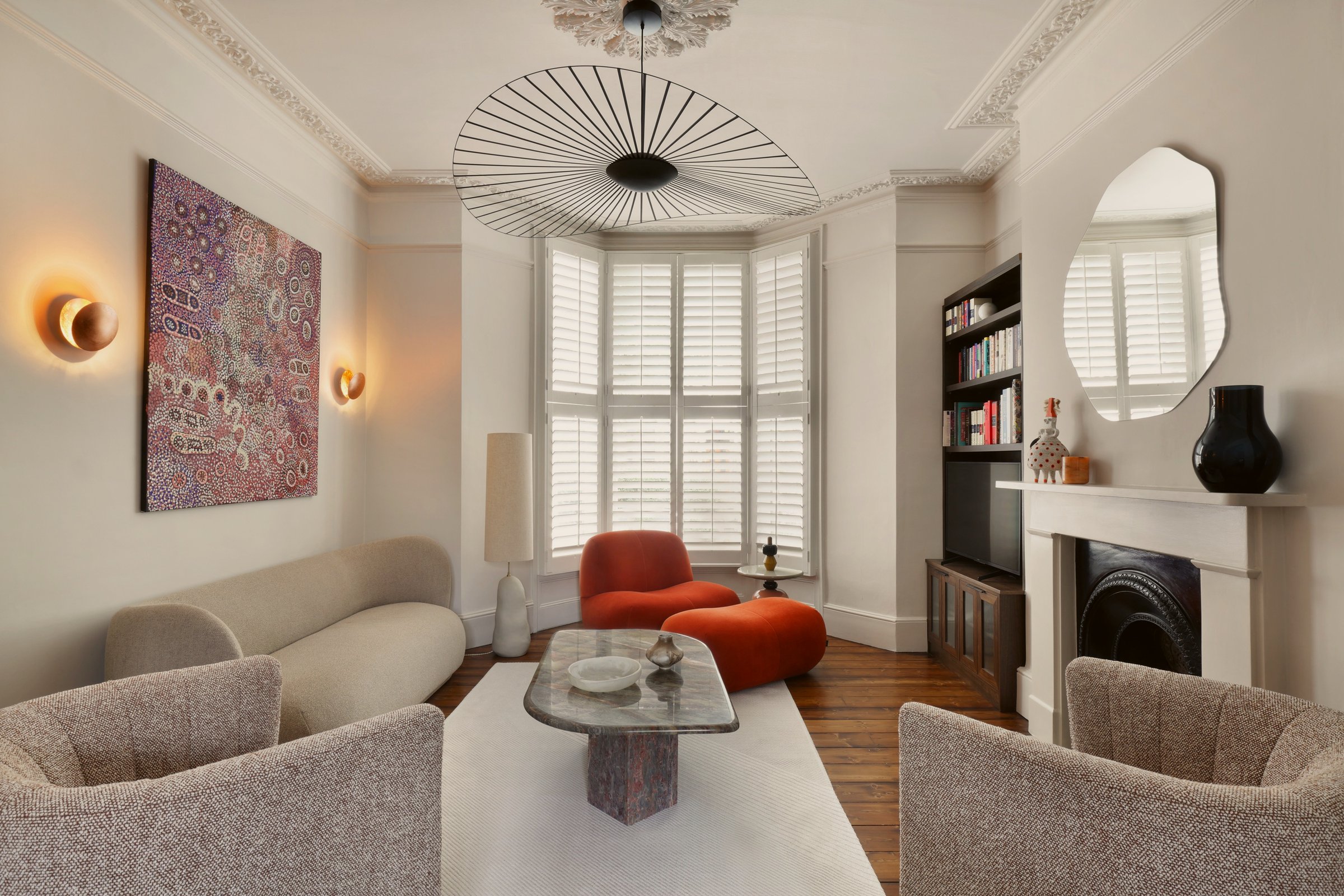 Sitting room with a shuttered bay window, a burnt orange armchair and footstool in the bay, a curved cream sofa and boucle tub chairs around a stone table.