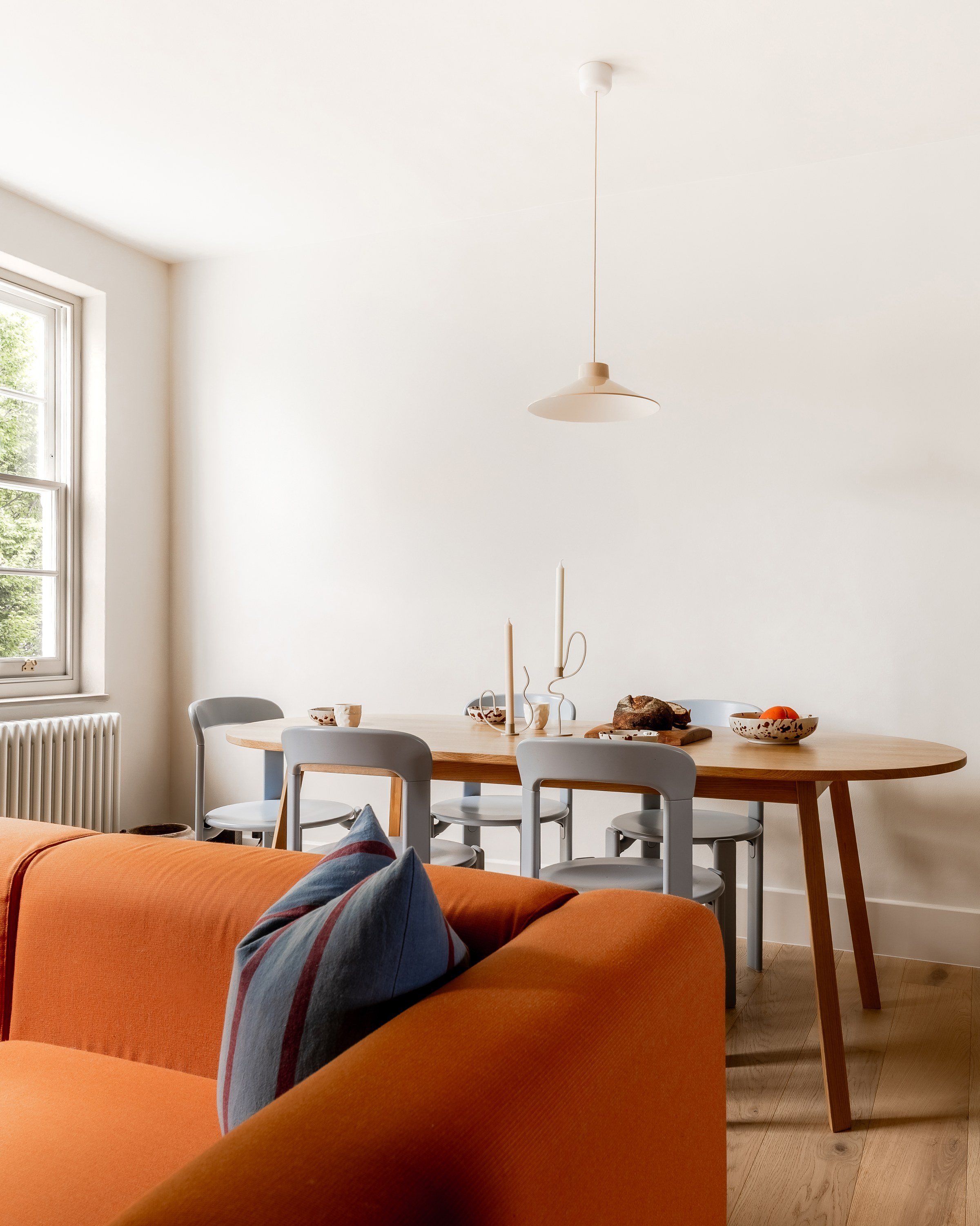Dining table with grey chairs under a pale pendant, seen past the arm of the orange sofa.