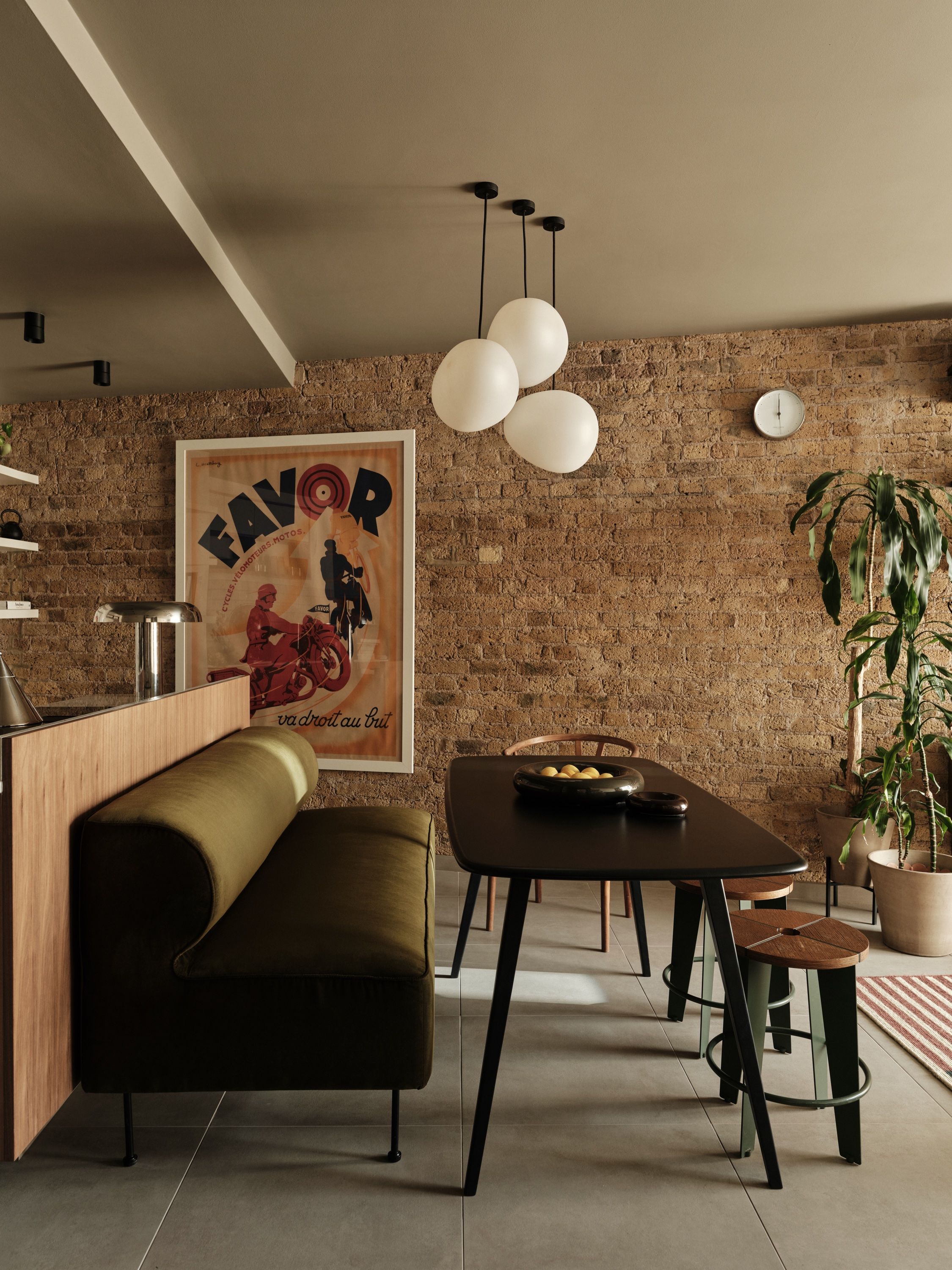 Dining area against exposed brick, with a dark table, an olive banquette built into the back of the kitchen counter and three white globe pendants.