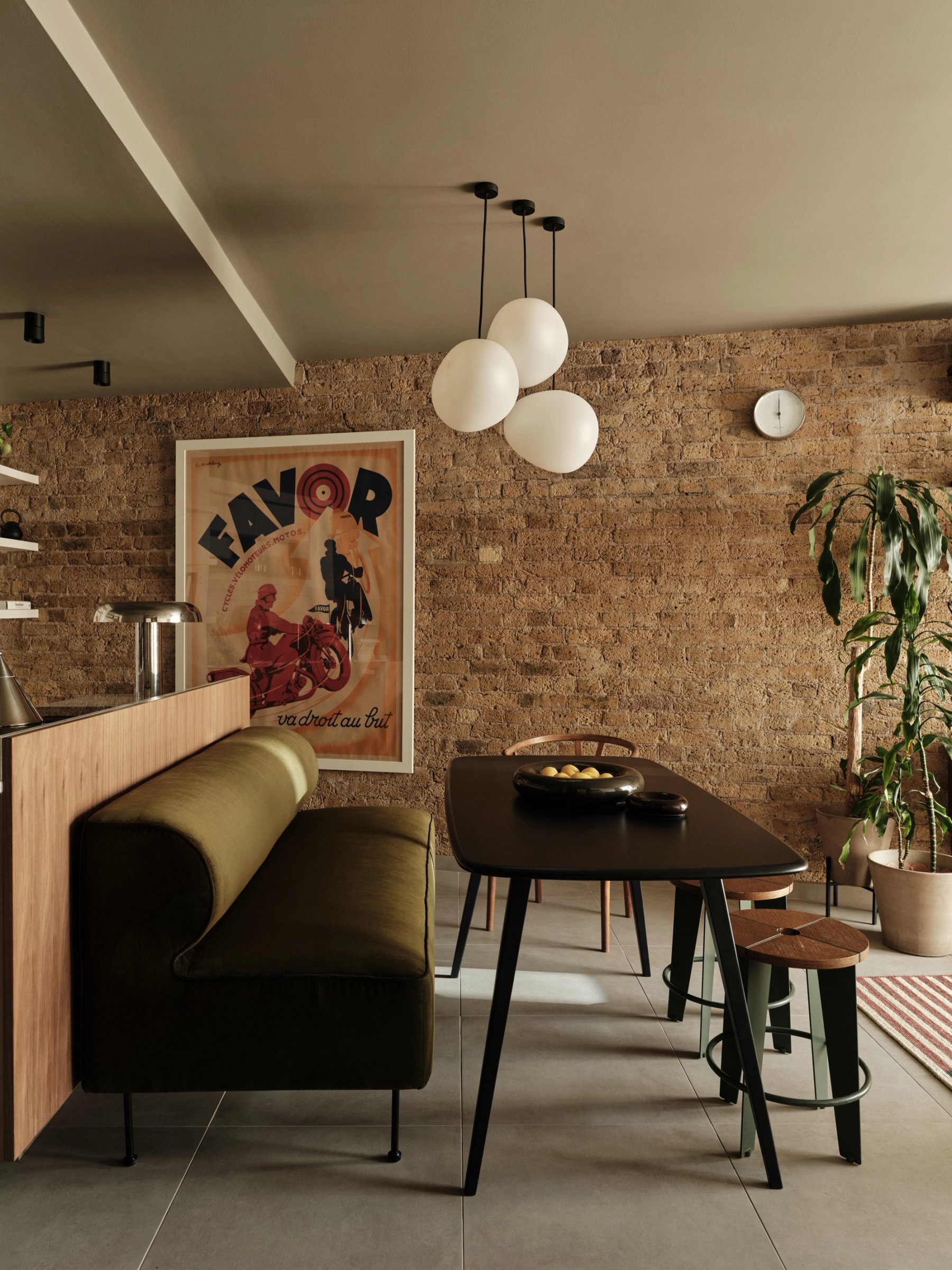Dining area against exposed brick, with a dark table, an olive banquette built into the back of the kitchen counter and three white globe pendants.