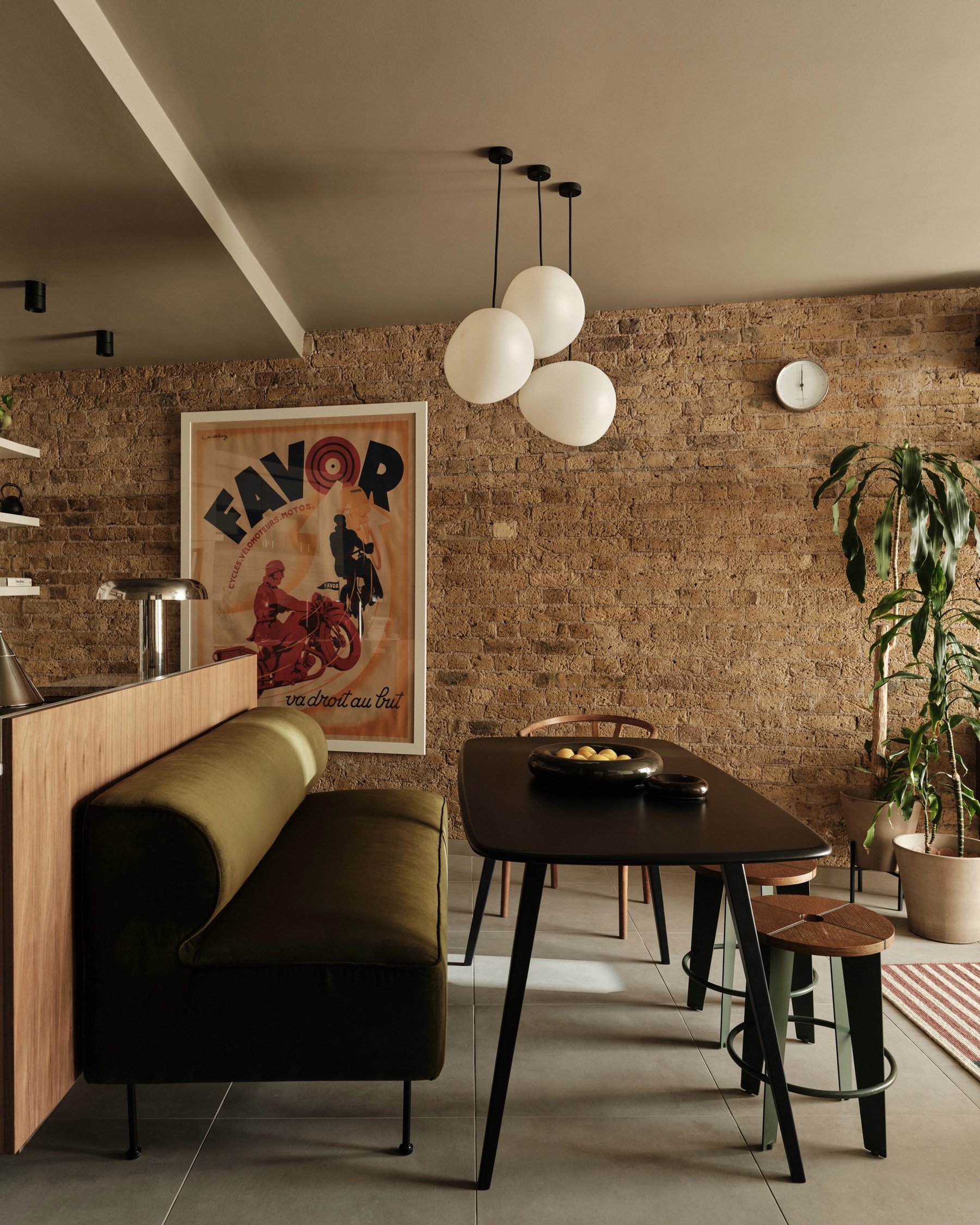 Dining area against exposed brick, with a dark table, an olive banquette built into the back of the kitchen counter and three white globe pendants.