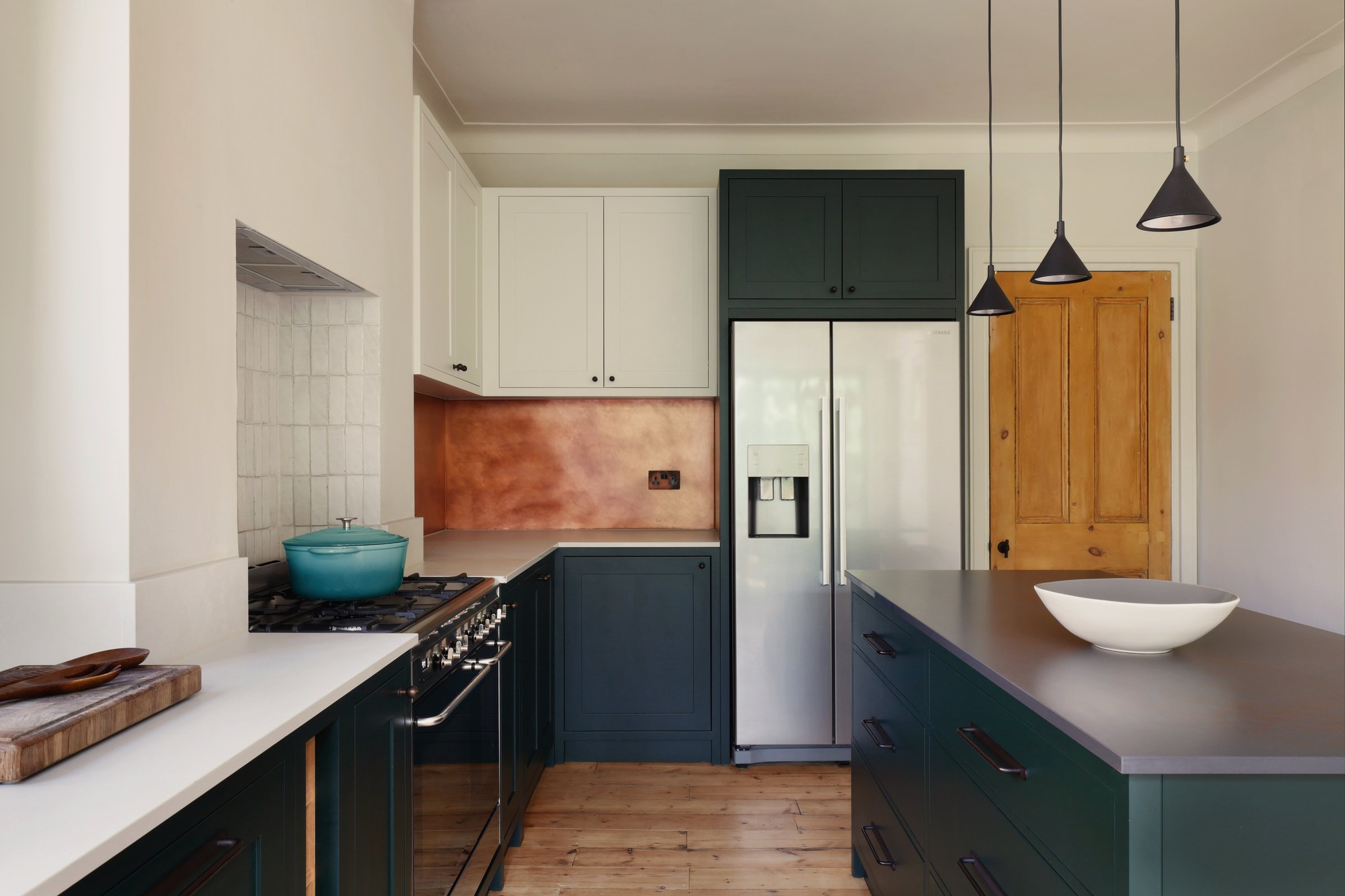 Kitchen seen across the island to a stripped pine door, with a copper splashback, a tall green cupboard for the fridge and three black cone pendants.