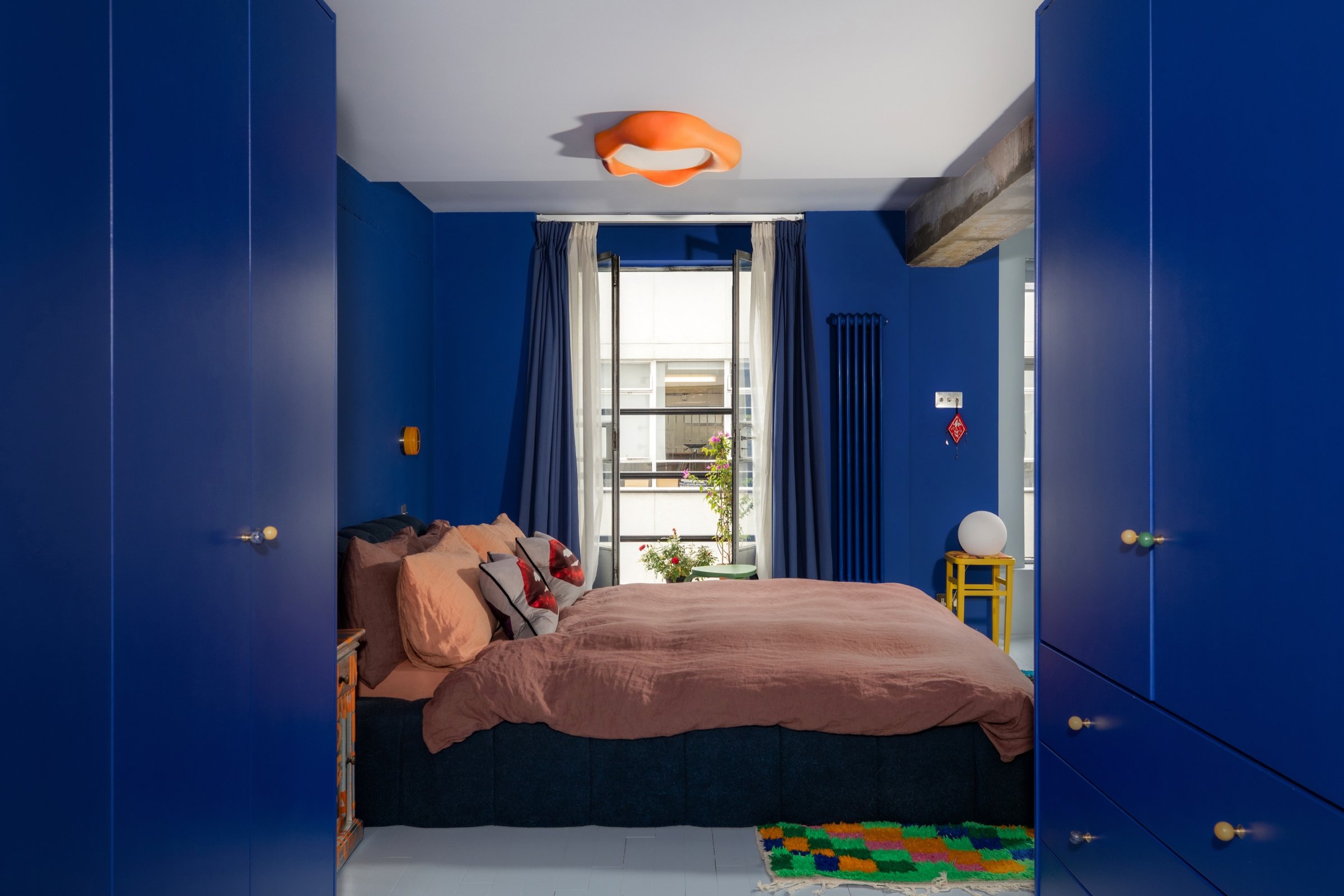 Blue bedroom framed symmetrically between two runs of deep blue fitted wardrobes, looking past the bed to the balcony door over a checkerboard rug.