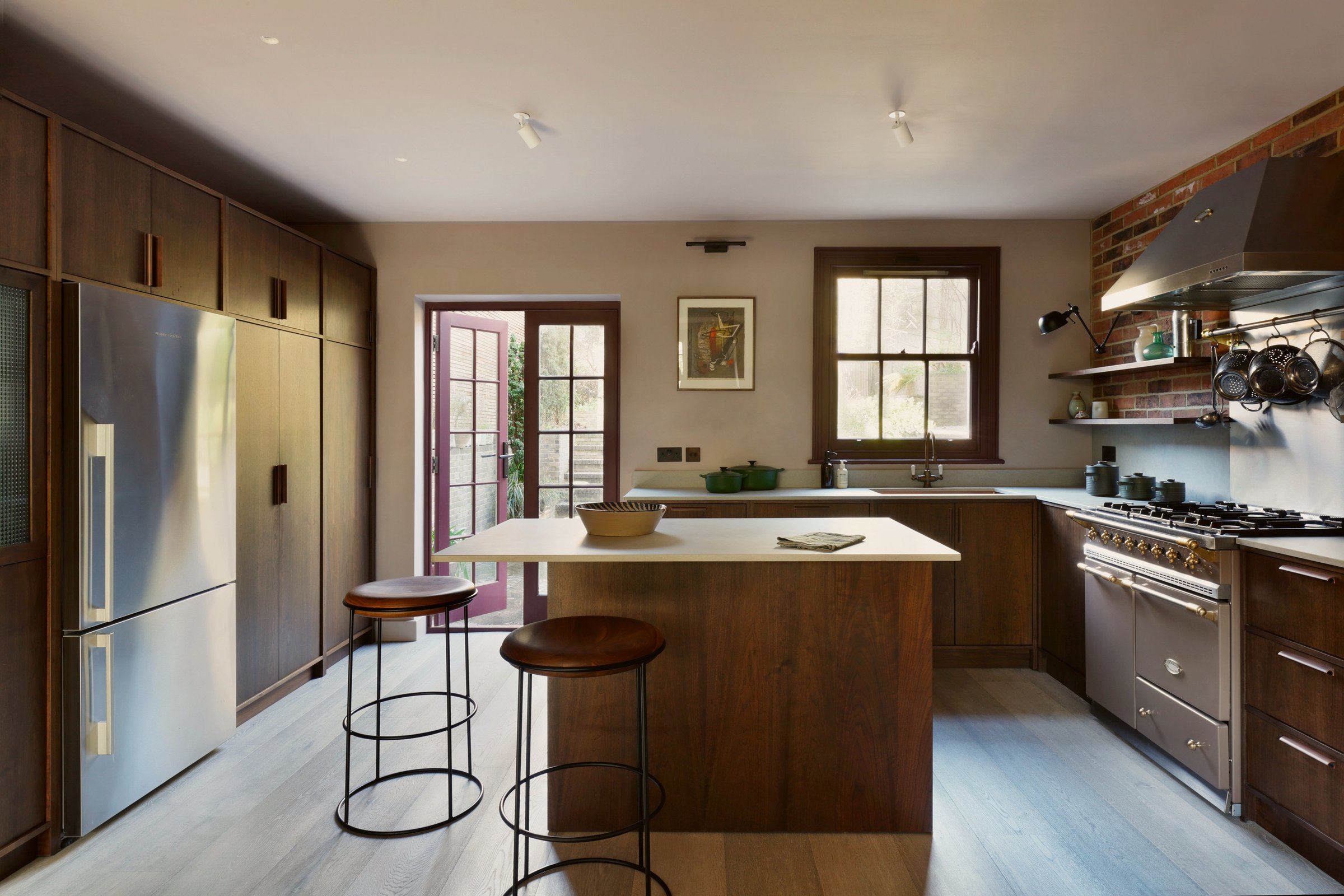 Kitchen across a dark timber island with two stools to glazed garden doors in a red-painted reveal, range cooker and brick chimney breast right.