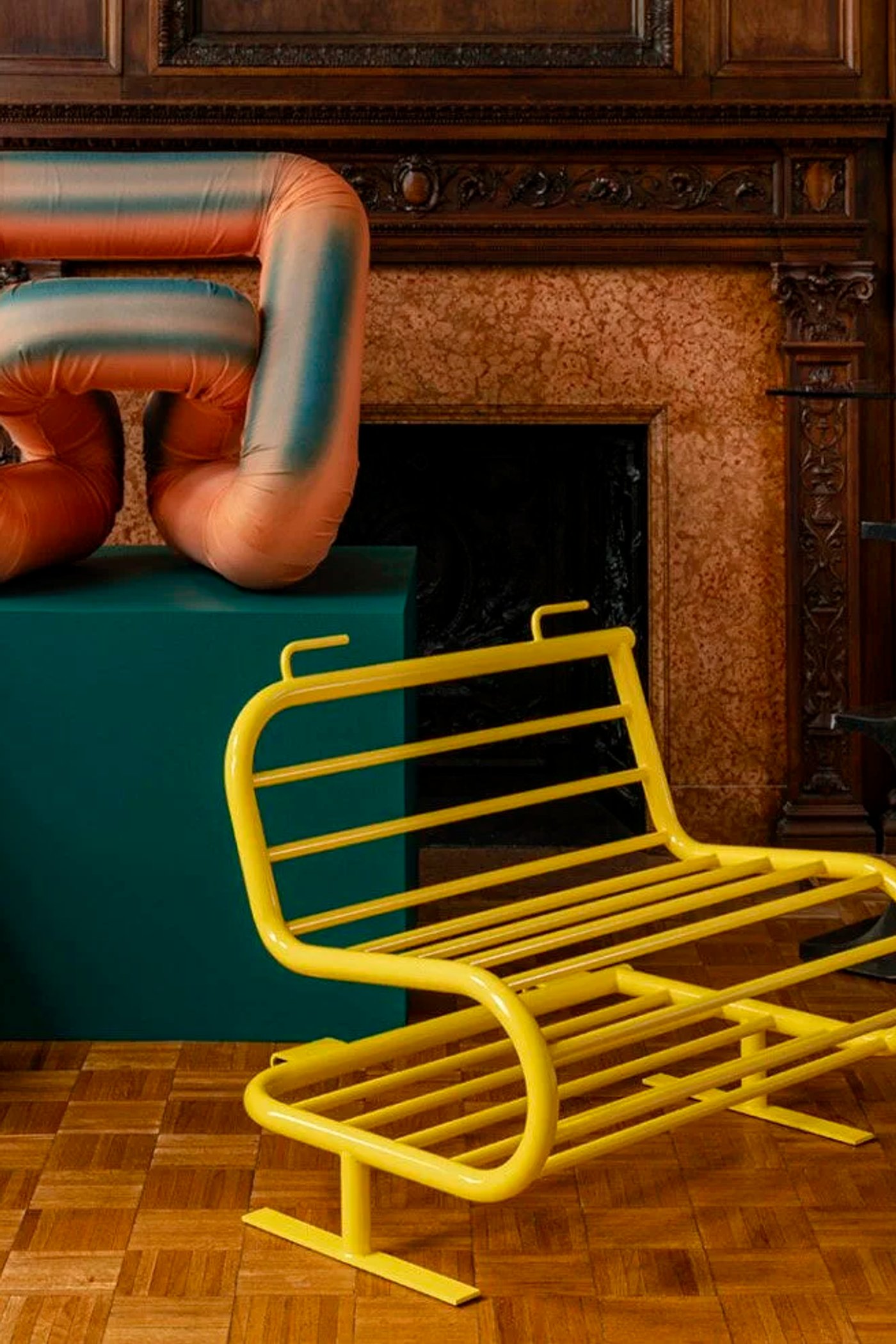 Panelled room with a carved stone fireplace, a yellow tubular steel bench, a padded terracotta form on a green plinth and a blue lattice stool