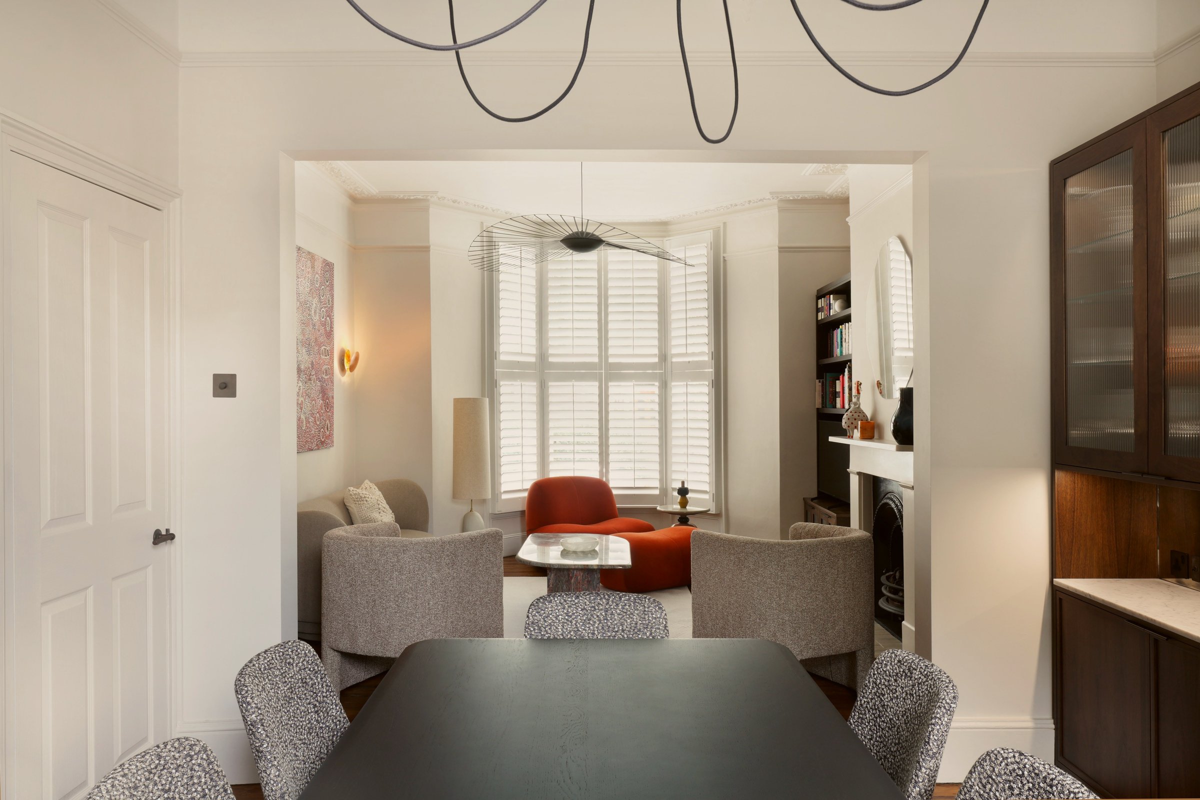 Dining room looking through a wide cornice-framed opening to the front sitting room, where a burnt orange armchair sits in a shuttered bay window.