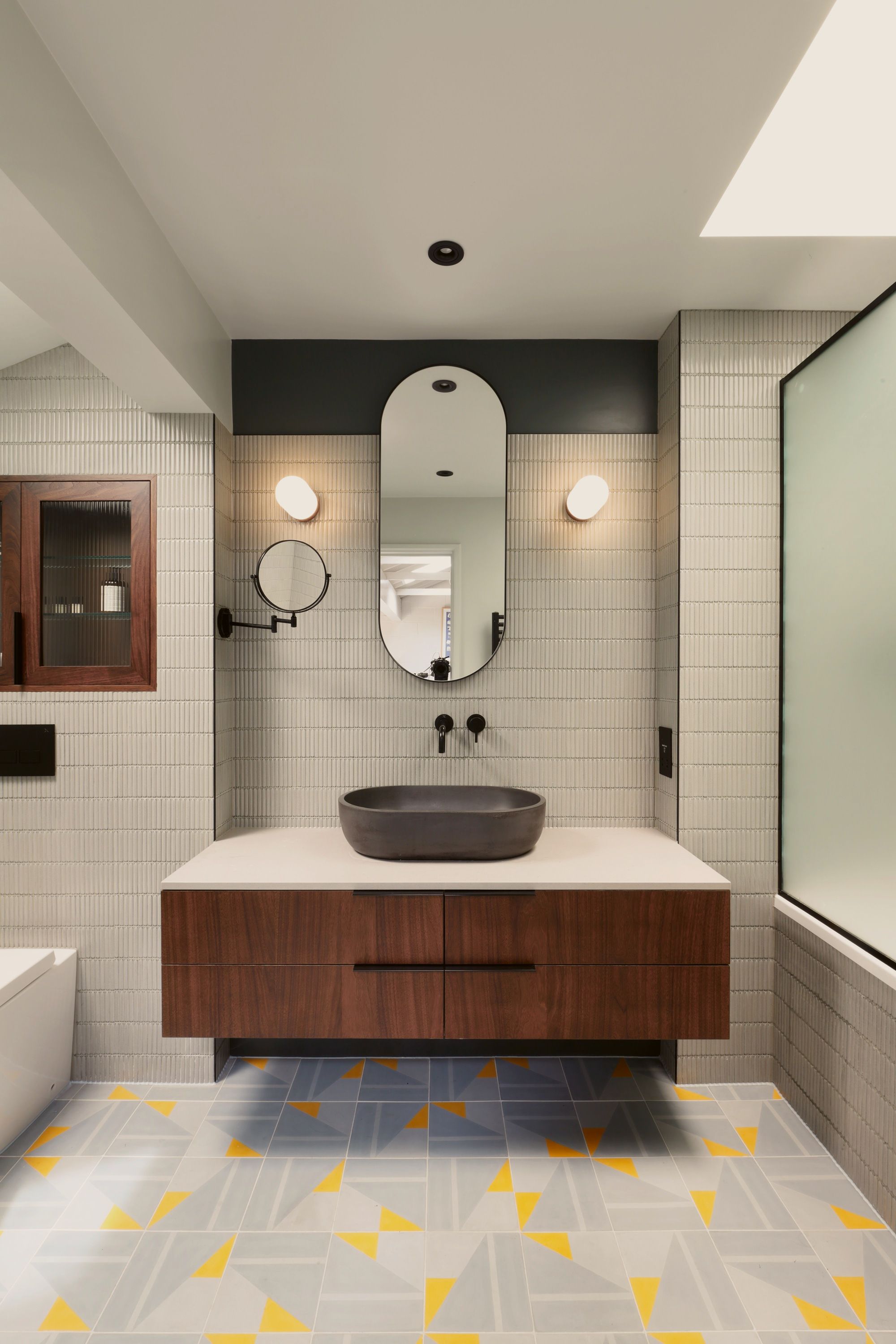 Bathroom with a walnut vanity floating below a pale stone top and dark stone basin, a capsule mirror between globe lights, on a yellow and grey tiled floor.