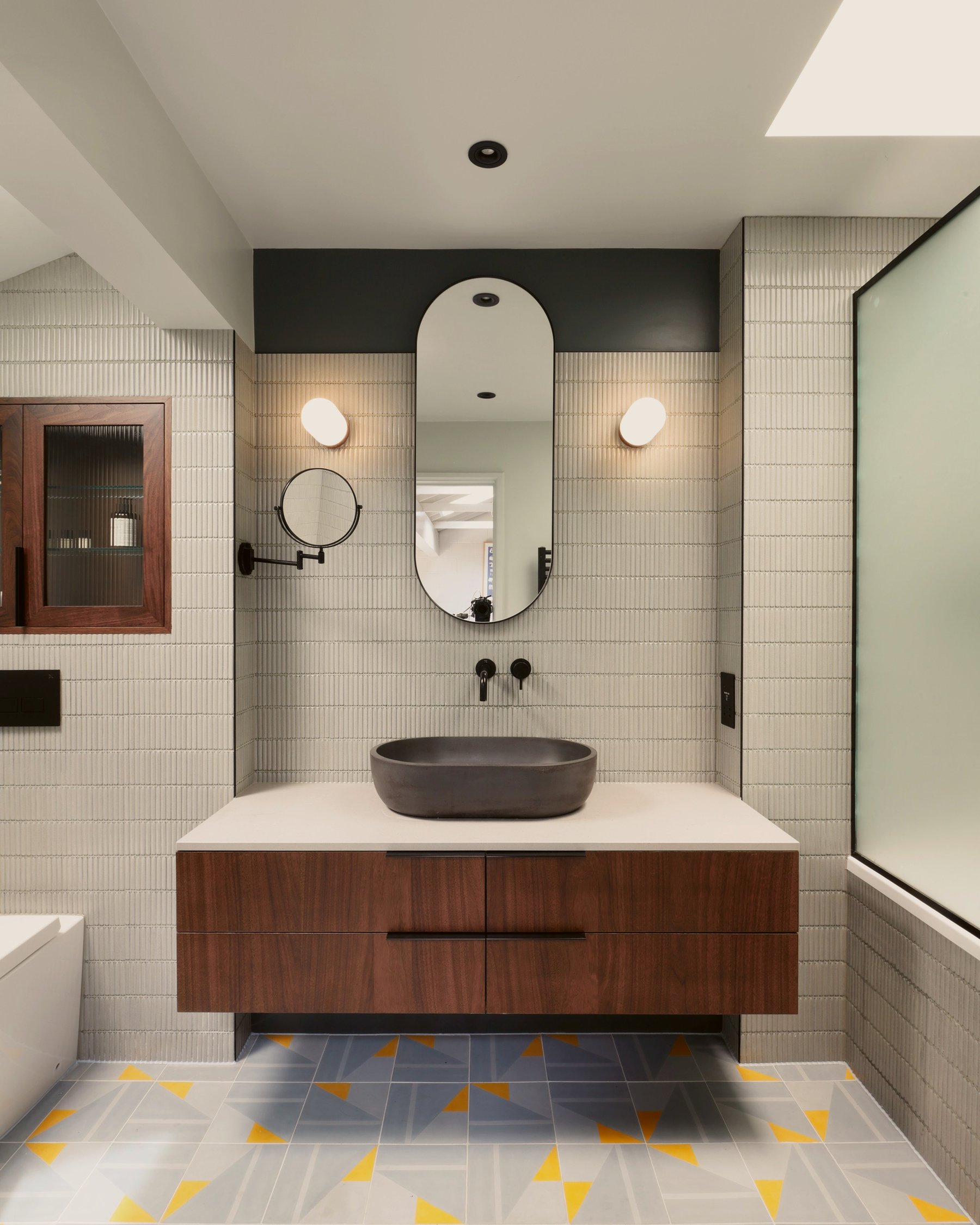 Bathroom with a walnut vanity floating below a pale stone top and dark stone basin, a capsule mirror between globe lights, on a yellow and grey tiled floor.