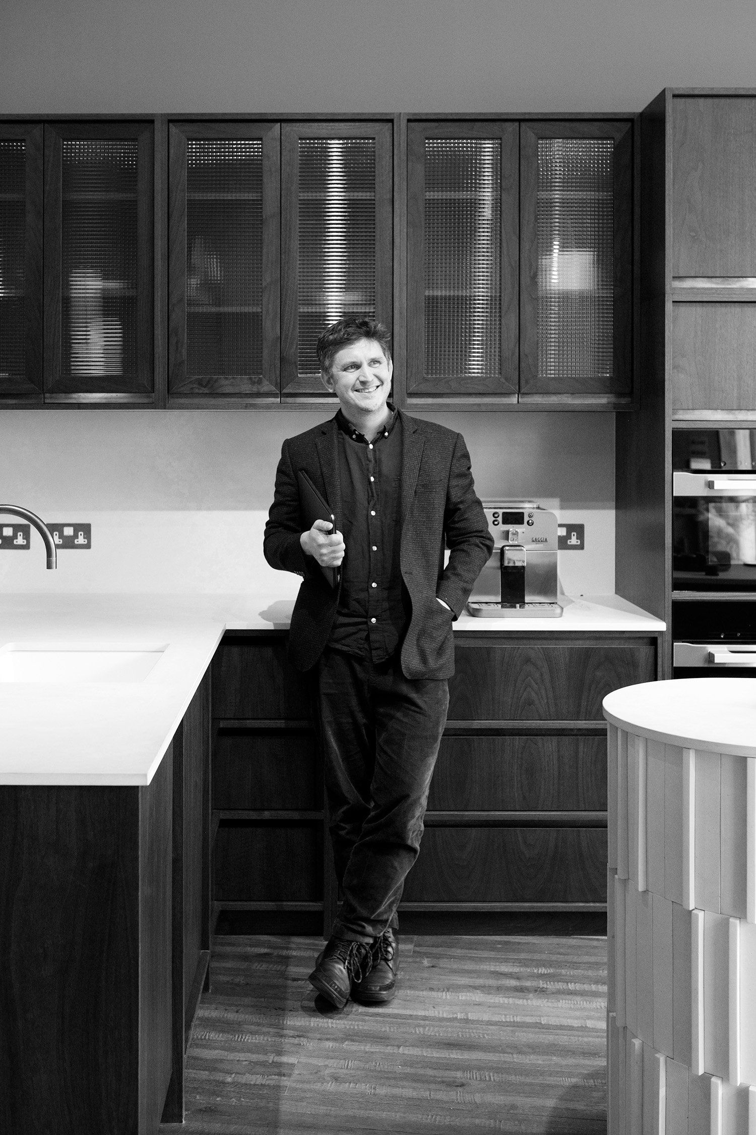 Luke Ellis leaning against a dark timber kitchen beneath reeded-glass wall cabinets, smiling and looking to one side.