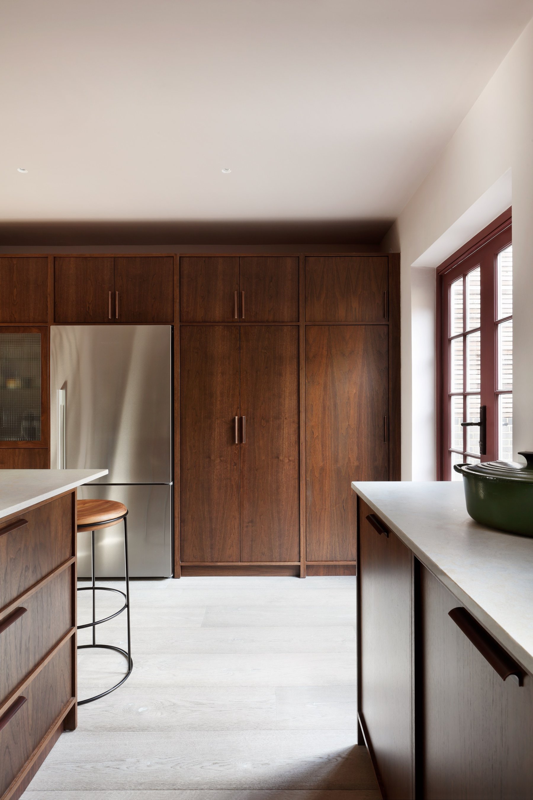 Dark stained tall cupboards beside an integrated fridge freezer, the island worktop in front and an arched red-framed window at the right.