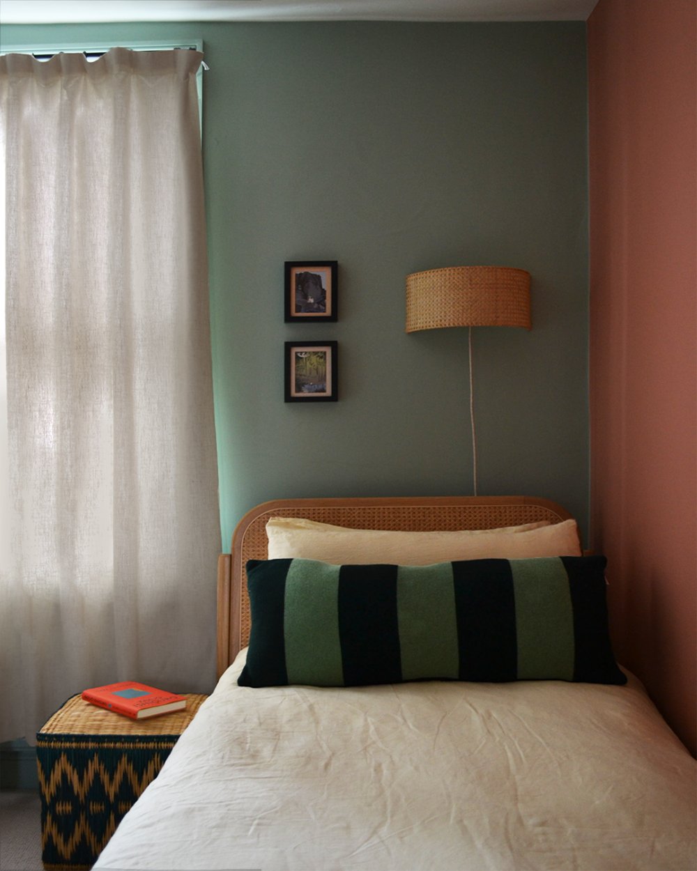 Bedroom with sage green and terracotta painted walls, a rattan headboard, striped bolster cushion and a woven wall light