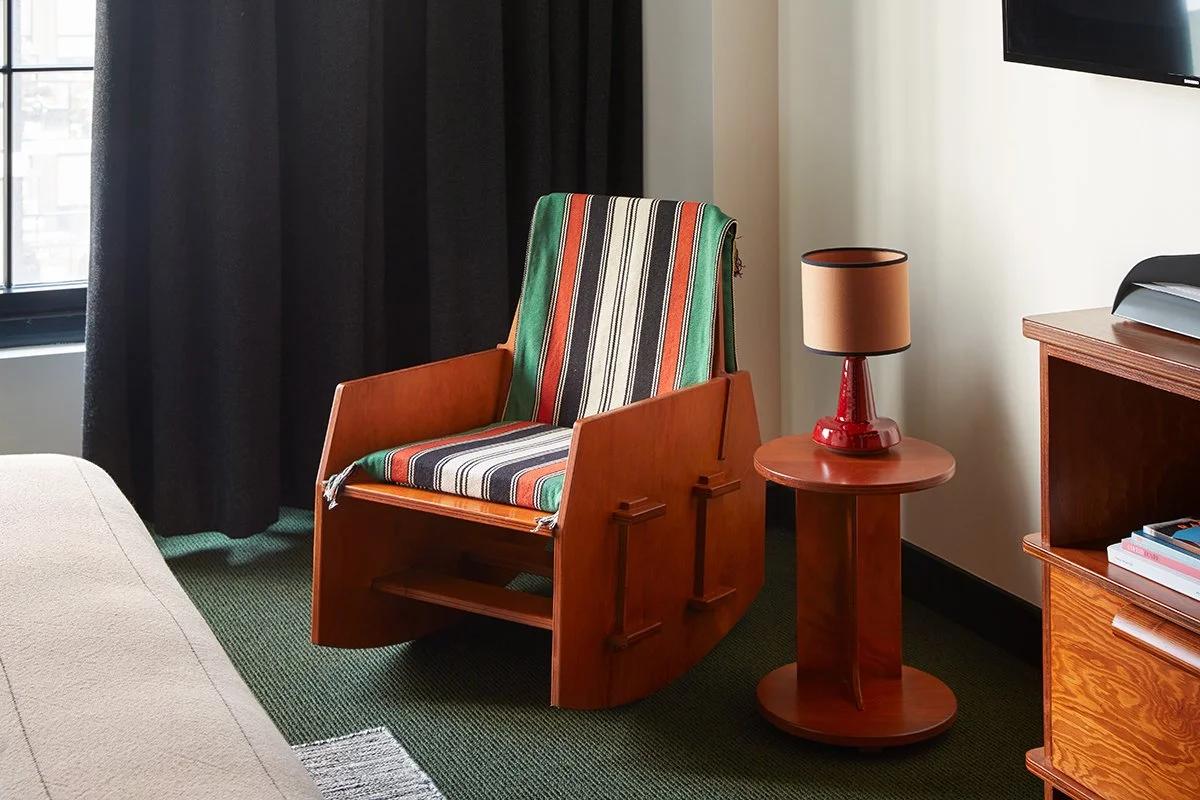 Hotel room corner with a cherry-red wooden rocking chair under a striped throw, beside a small red side table and lamp on green carpet